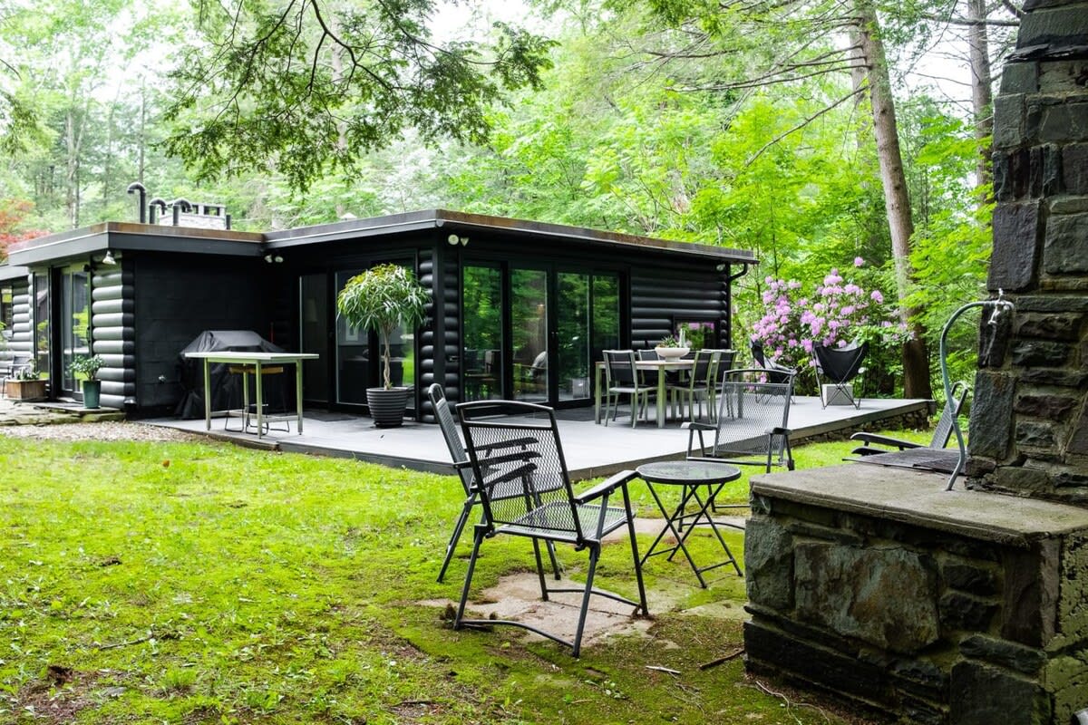 The back exterior of a modern black log cabin, featuring a large gray deck with an outdoor dining set and lounge chairs, surrounded by lush green trees and grass. A stone outdoor fireplace is visible on the right.