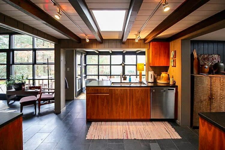 A mid-century modern kitchen with dark wood cabinetry, black countertops, and a stainless steel dishwasher, illuminated by track lighting and a skylight. A small rug is on the dark tiled floor.