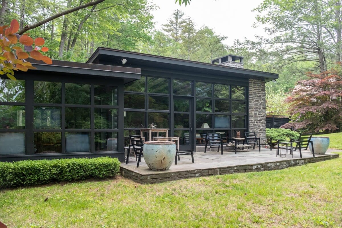 A modern cabin with a dark exterior, featuring a wall of large black-framed windows and a stone patio with outdoor seating. Lush green trees surround the property, and a manicured hedge borders the patio.