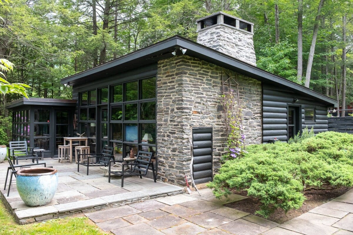 The side and back of a modern cabin, showcasing a striking combination of dark stone masonry and black horizontal log siding, with a prominent stone chimney. A flagstone patio with outdoor seating is visible, surrounded by dense forest.