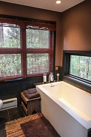 A modern bathroom with dark brown walls and dark tiled floors, featuring a deep white soaking tub next to a large window with bamboo blinds. A small wooden stool with towels sits beside the tub.