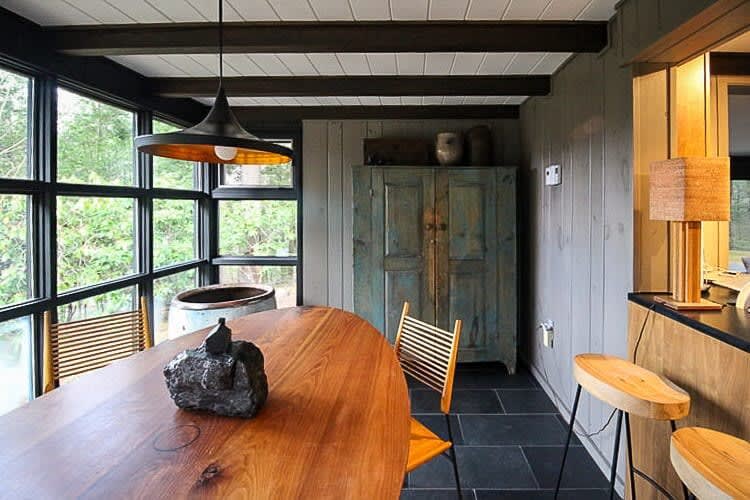 A dining area with a large oval wooden table and two slatted wooden chairs, surrounded by floor-to-ceiling windows overlooking a forest. A dark pendant light hangs above the table, and an antique blue cabinet stands against a paneled wall.