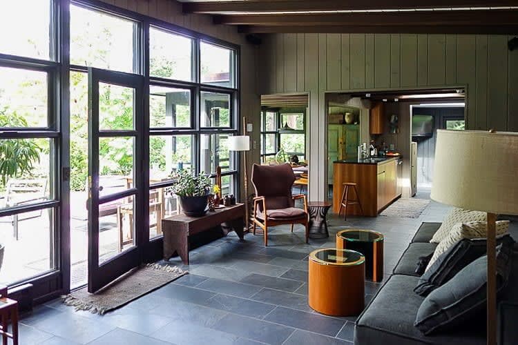 A bright living space with dark slate tile floors and expansive black-framed windows leading to an outdoor patio. A comfortable armchair, wooden bench, and a glimpse of the kitchen with a wooden island are visible, all under exposed ceiling beams.