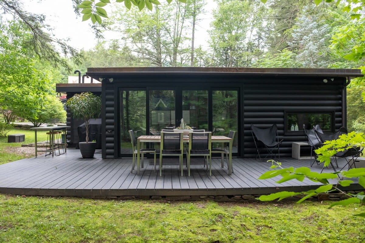 A wider view of the modern black log cabin's back exterior, showcasing a spacious gray deck with an outdoor dining table and chairs, and large sliding glass doors. The cabin is nestled among green trees and a grassy yard.