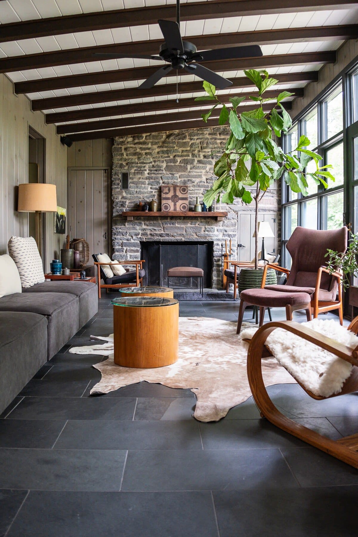 A spacious living room with dark slate tile floors, exposed wooden beams, and a large stone fireplace. A long grey sofa, a cowhide rug, and various mid-century modern chairs create a comfortable and stylish atmosphere with ample natural light.