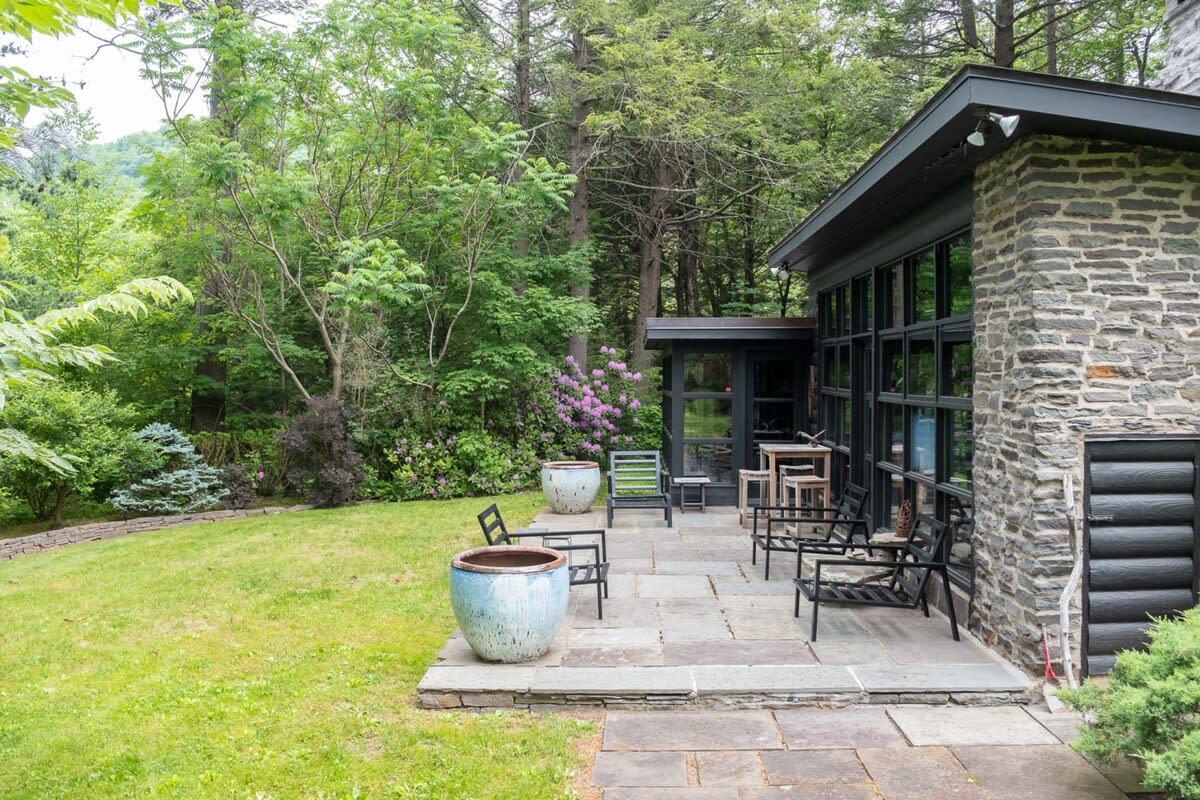 A wide view of the cabin's flagstone patio, furnished with black outdoor chairs and large ceramic planters, extending into a grassy yard. The property is nestled among a vibrant green forest with flowering rhododendrons.
