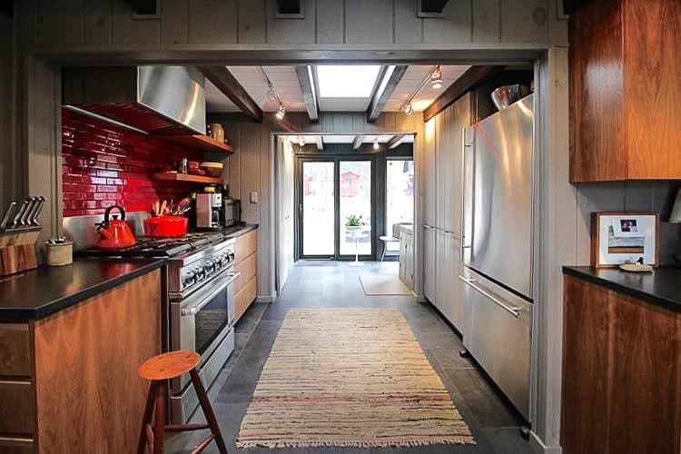 A galley kitchen featuring dark wood cabinetry, stainless steel appliances including a large refrigerator and range, and a vibrant red subway tile backsplash. A long woven rug runs down the center of the dark tiled floor.