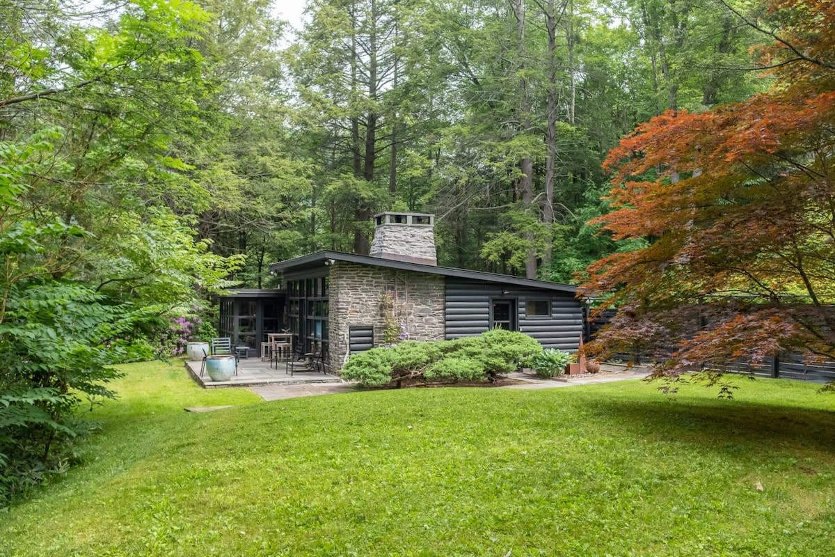 A full exterior view of the modern cabin, featuring a dark facade with stone and log siding, and a large stone chimney, set within a sprawling green lawn. The property is enveloped by a dense, lush forest, with a vibrant red-leafed tree in the foreground.