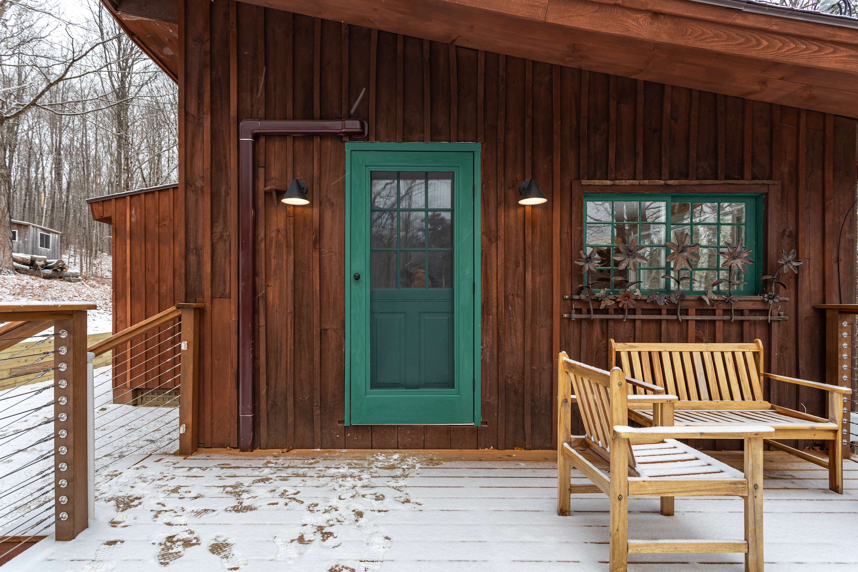 A close-up of the cabin's dark brown wooden exterior with a vibrant green entrance door and matching window frame, leading to a snow-dusted wooden deck with outdoor seating.