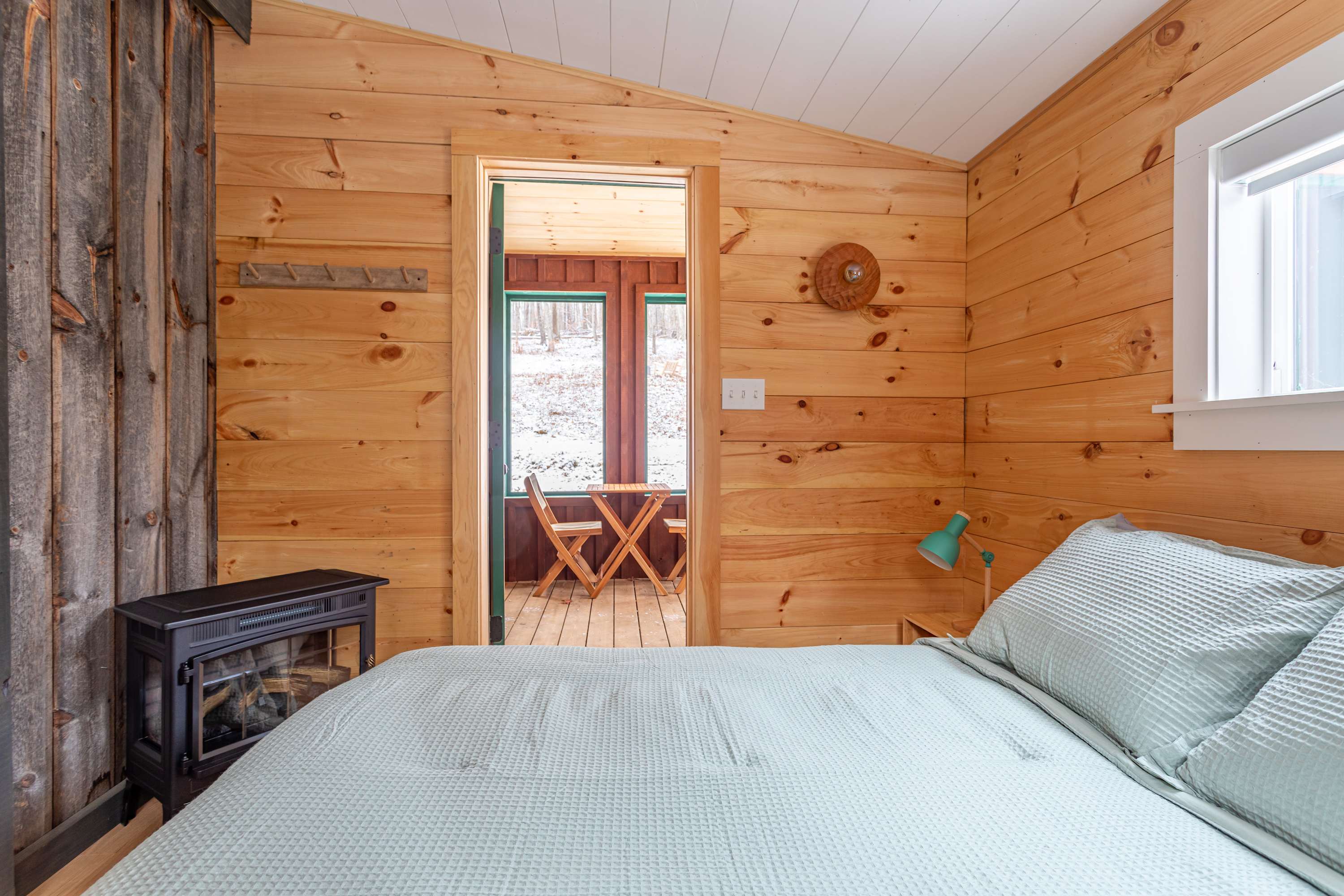Another view of the cozy bedroom with light wood paneling, showing a queen bed and a black electric fireplace in the corner. A doorway leads to an enclosed porch with outdoor seating visible through the window.