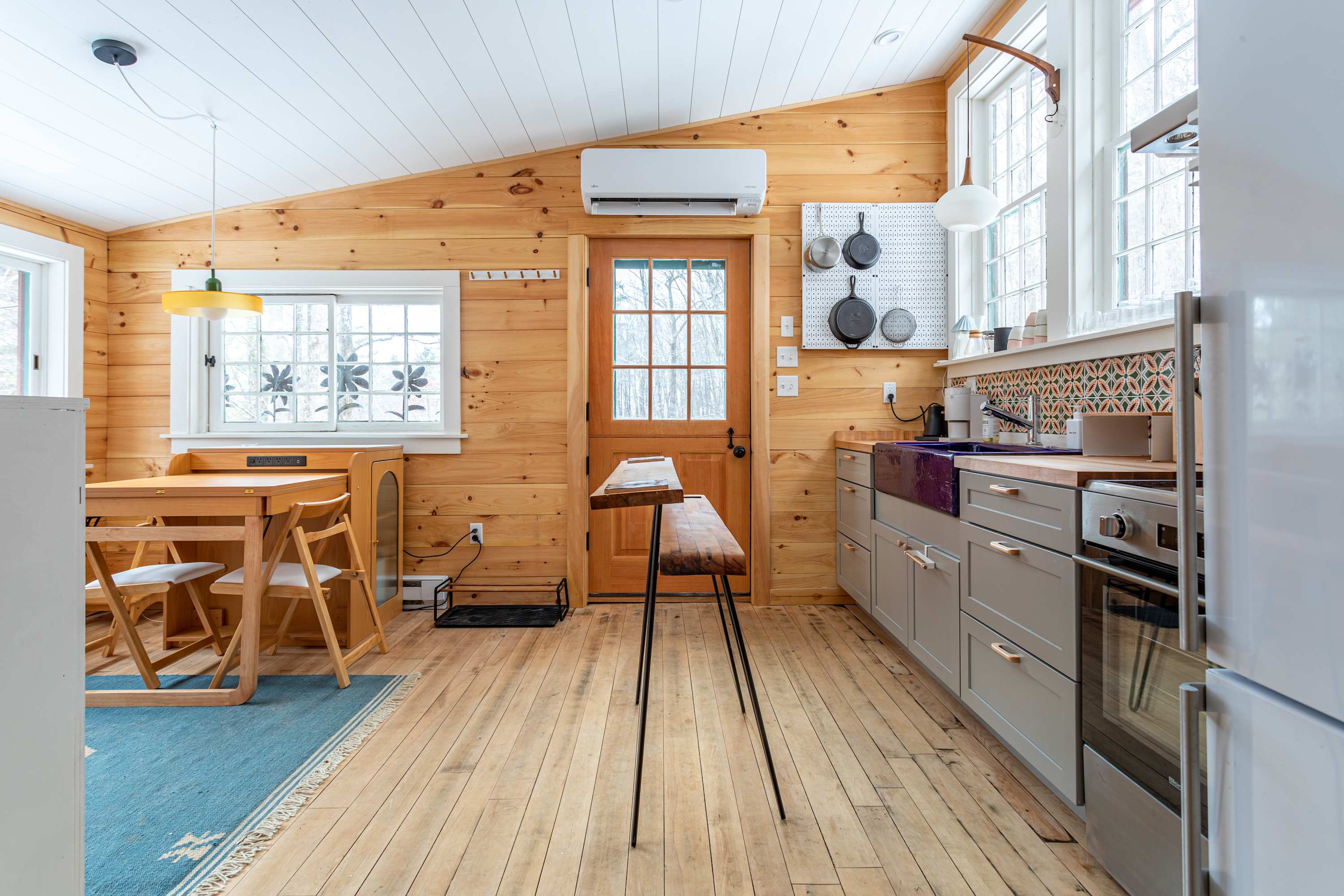 A bright kitchen and dining area with light wood plank walls and floor, featuring a small wooden dining table with chairs on a blue rug, and the kitchen counter with gray cabinets and a purple sink visible.