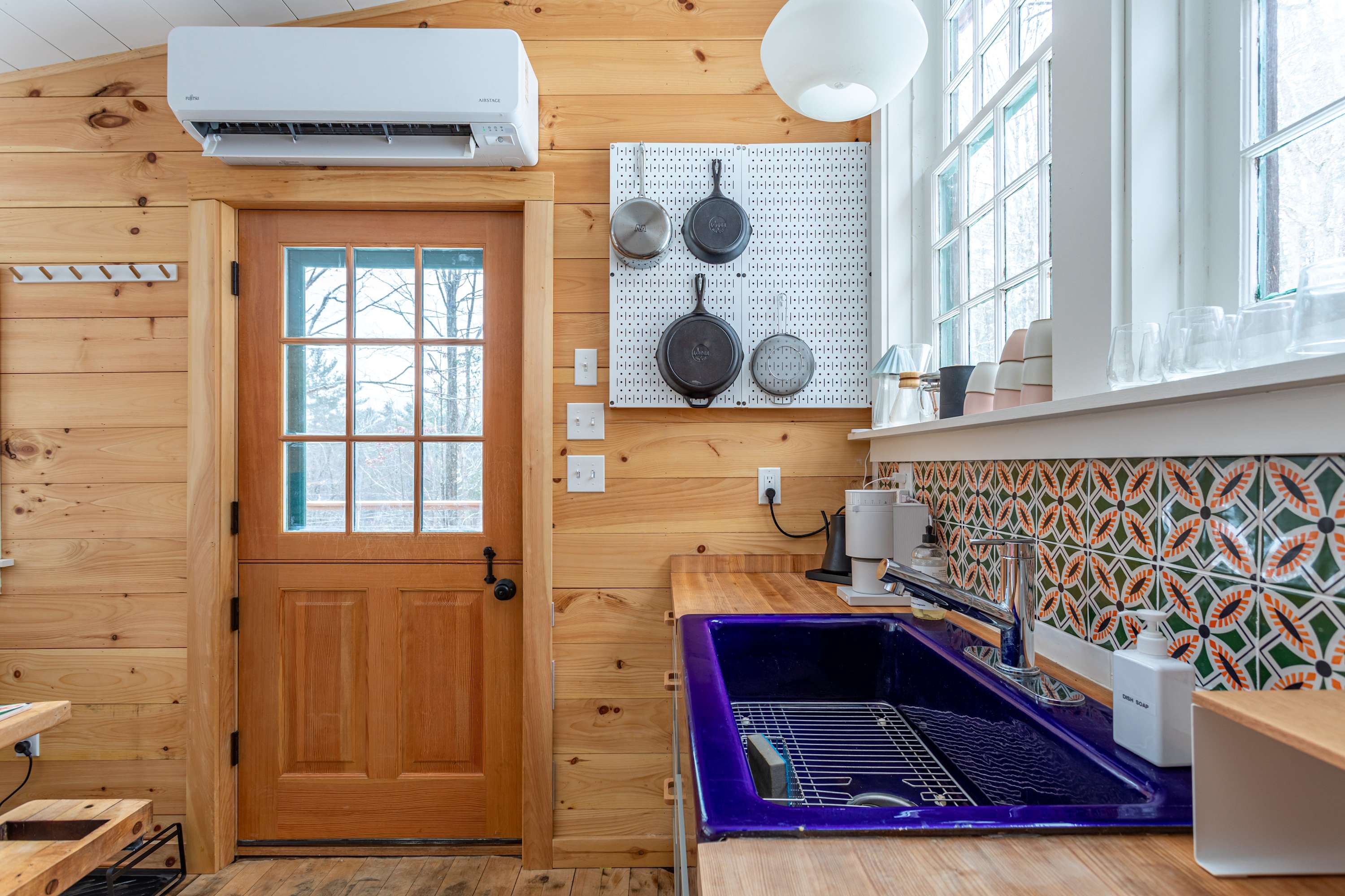 A rustic kitchen with light wood paneling, a vibrant purple sink, and a white pegboard displaying cast iron pans. A glass-paned door leads outside, and a white mini-split AC unit is mounted above.