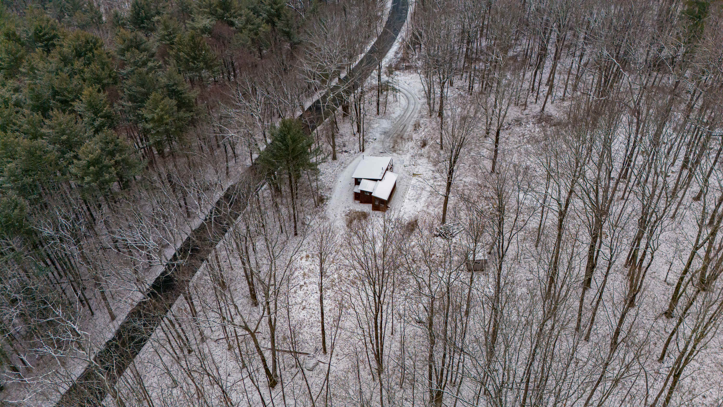 High aerial view of the cabin nestled in a snow-covered forest, with a winding road and a dark river visible, highlighting the property's remote and natural surroundings.