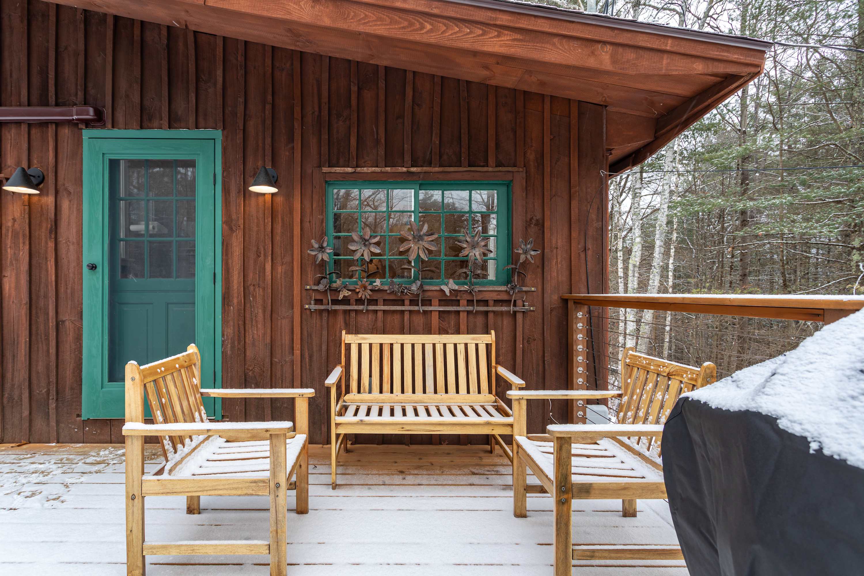 Outdoor wooden deck with a teal door and window, featuring a wooden bench and two chairs, all lightly dusted with snow, set against a backdrop of a snowy forest.