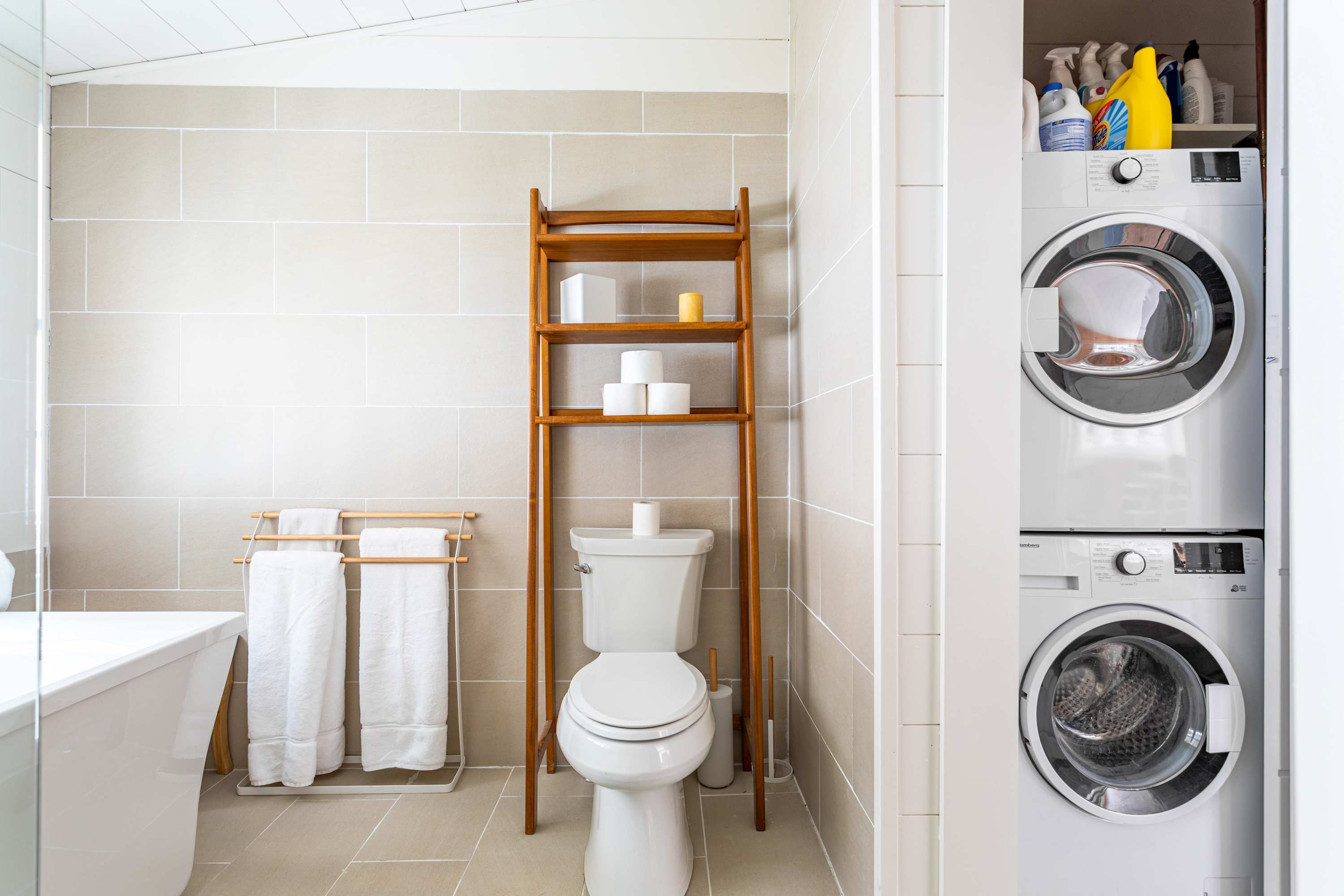 A bright bathroom with light beige tiled walls and floor, featuring a white toilet and a freestanding bathtub. A wooden ladder shelf provides storage, and a stacked washer and dryer unit is visible in an adjacent closet.