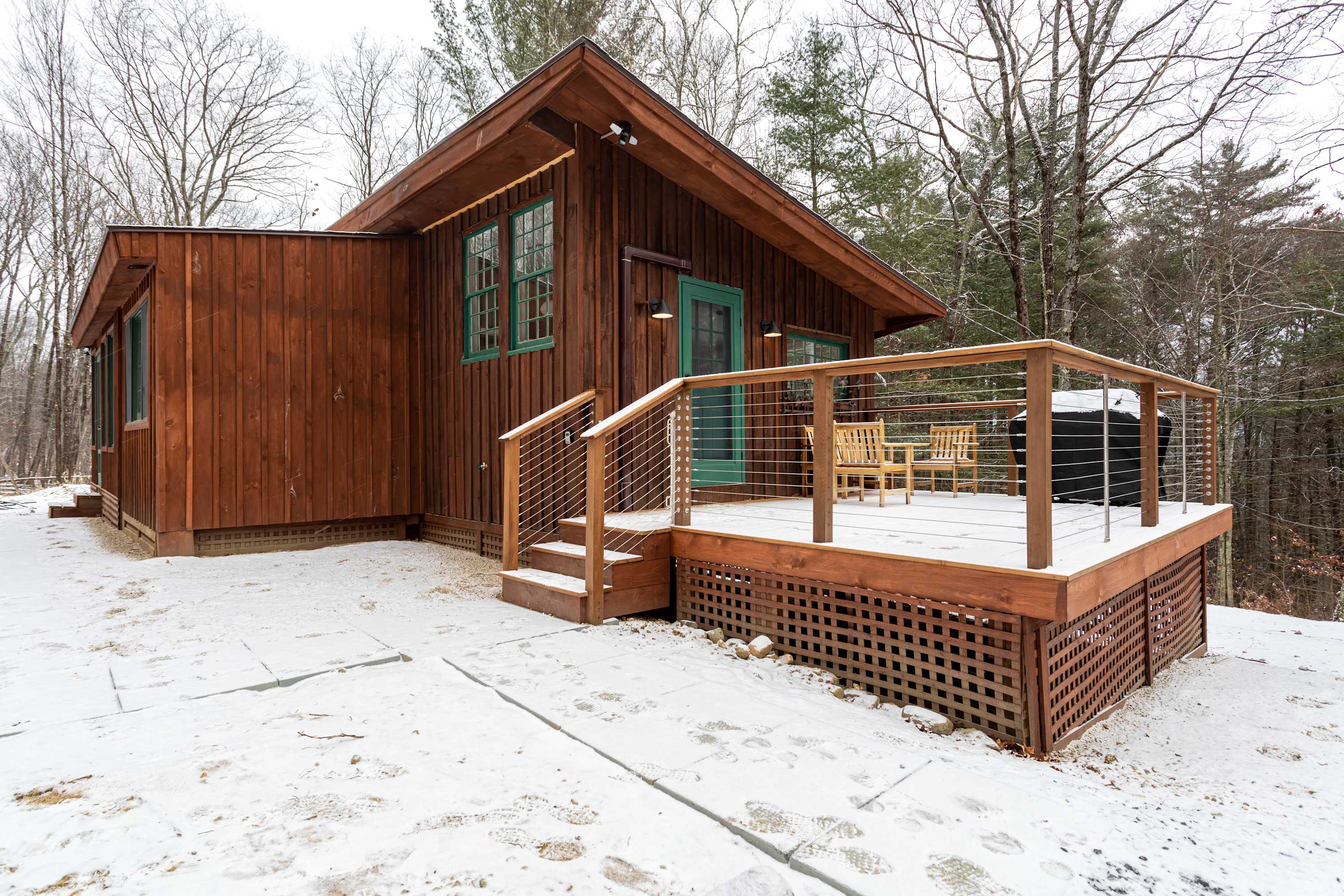 Charming brown wooden cabin with a sloped roof and a front deck, partially covered in snow, featuring teal-trimmed windows and doors, nestled in a snowy, wooded landscape.
