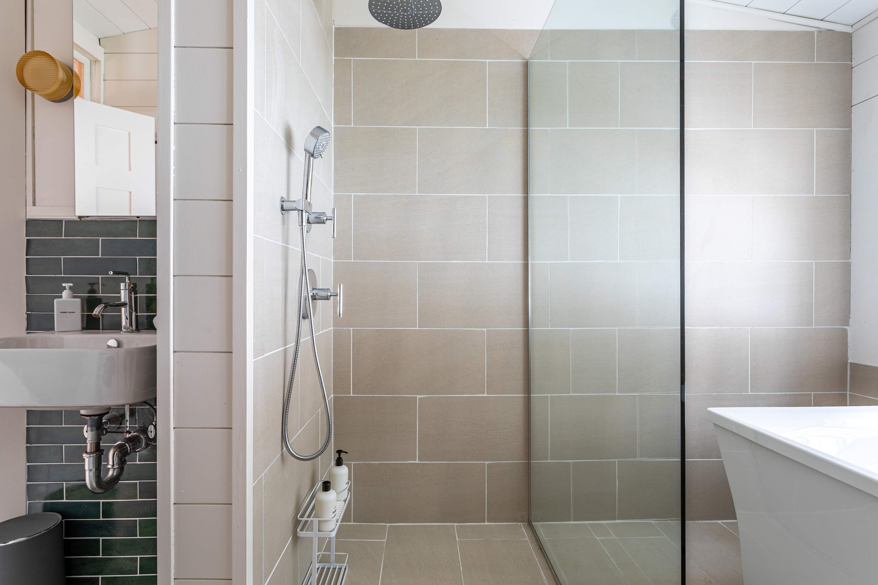 A modern bathroom featuring a spacious walk-in shower with light beige subway tiles and a glass partition. A small wall-mounted sink with a dark green tiled backsplash is visible to the left.