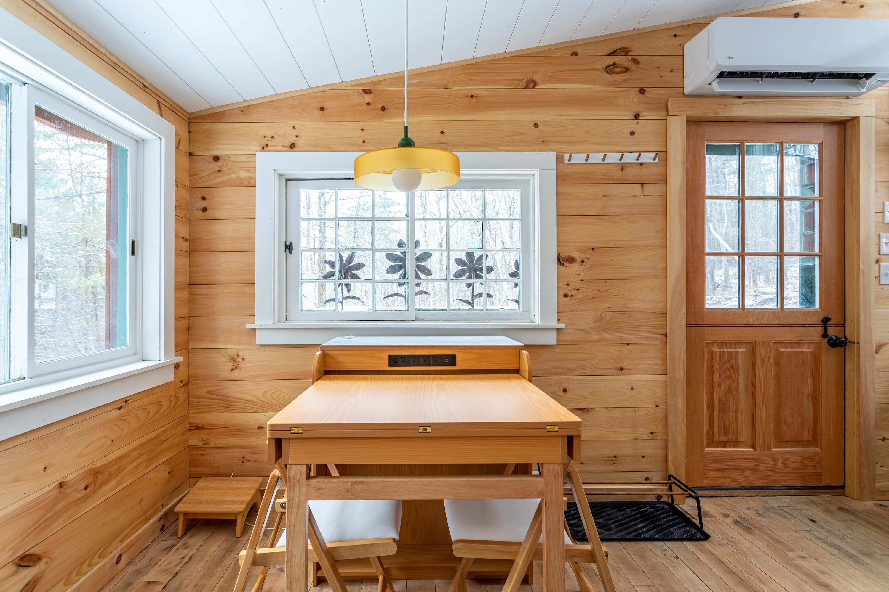 A cozy dining nook with light wood paneling and a sloped white ceiling, featuring a compact wooden table with built-in seating and a yellow pendant light. A glass-paned door and windows provide natural light.