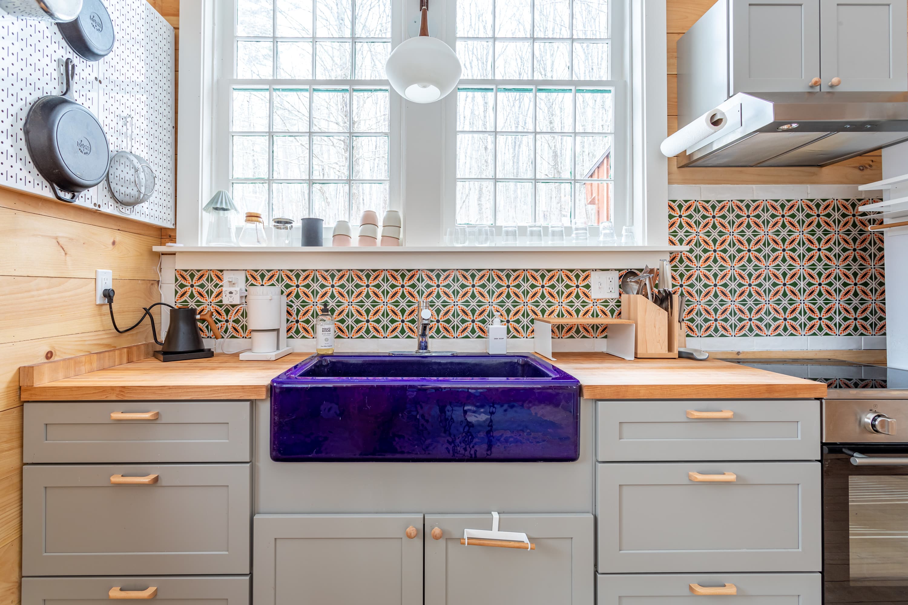 A detailed view of the kitchen's vibrant purple farmhouse sink set into butcher block countertops, complemented by light gray cabinets and a striking geometric tile backsplash, with light wood plank walls.