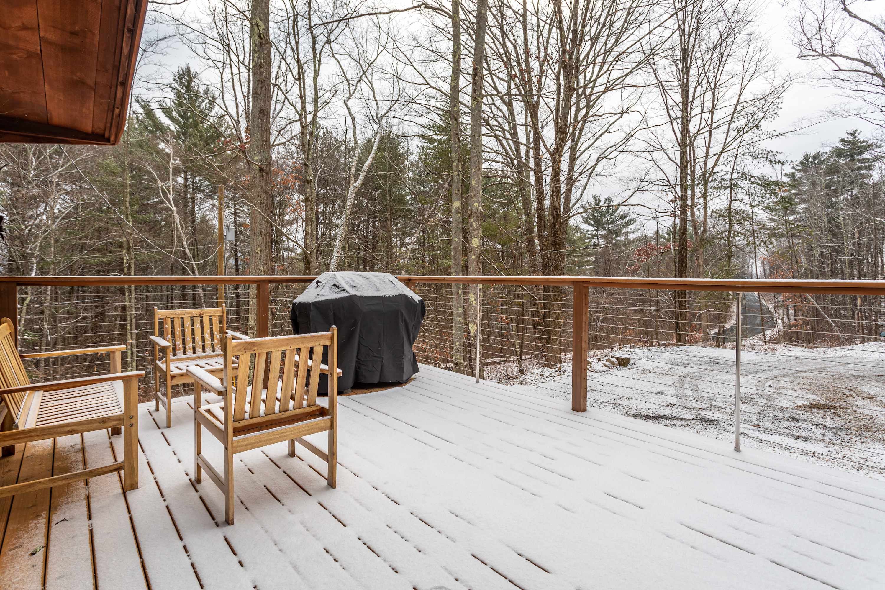 Expansive wooden deck covered in a light layer of snow, featuring a black BBQ grill and several wooden chairs, with a cable railing overlooking a snowy forest and a distant road.