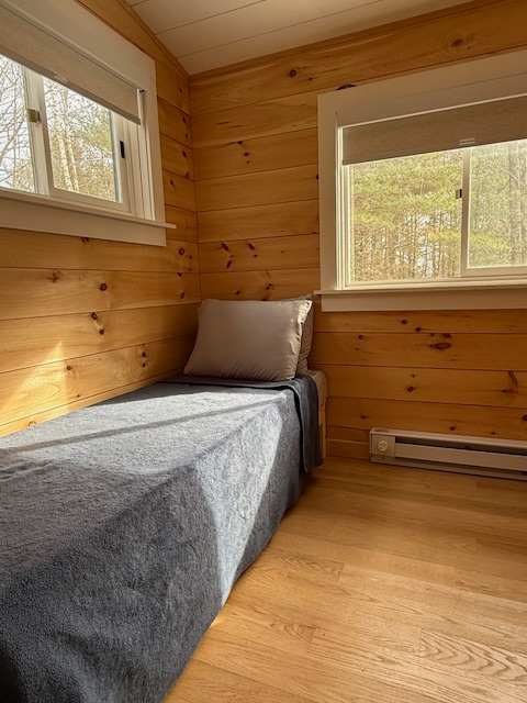 Bright bedroom with light wood paneled walls and hardwood floors, featuring a single bed with a grey blanket and pillow, and two windows offering views of a sunlit forest.