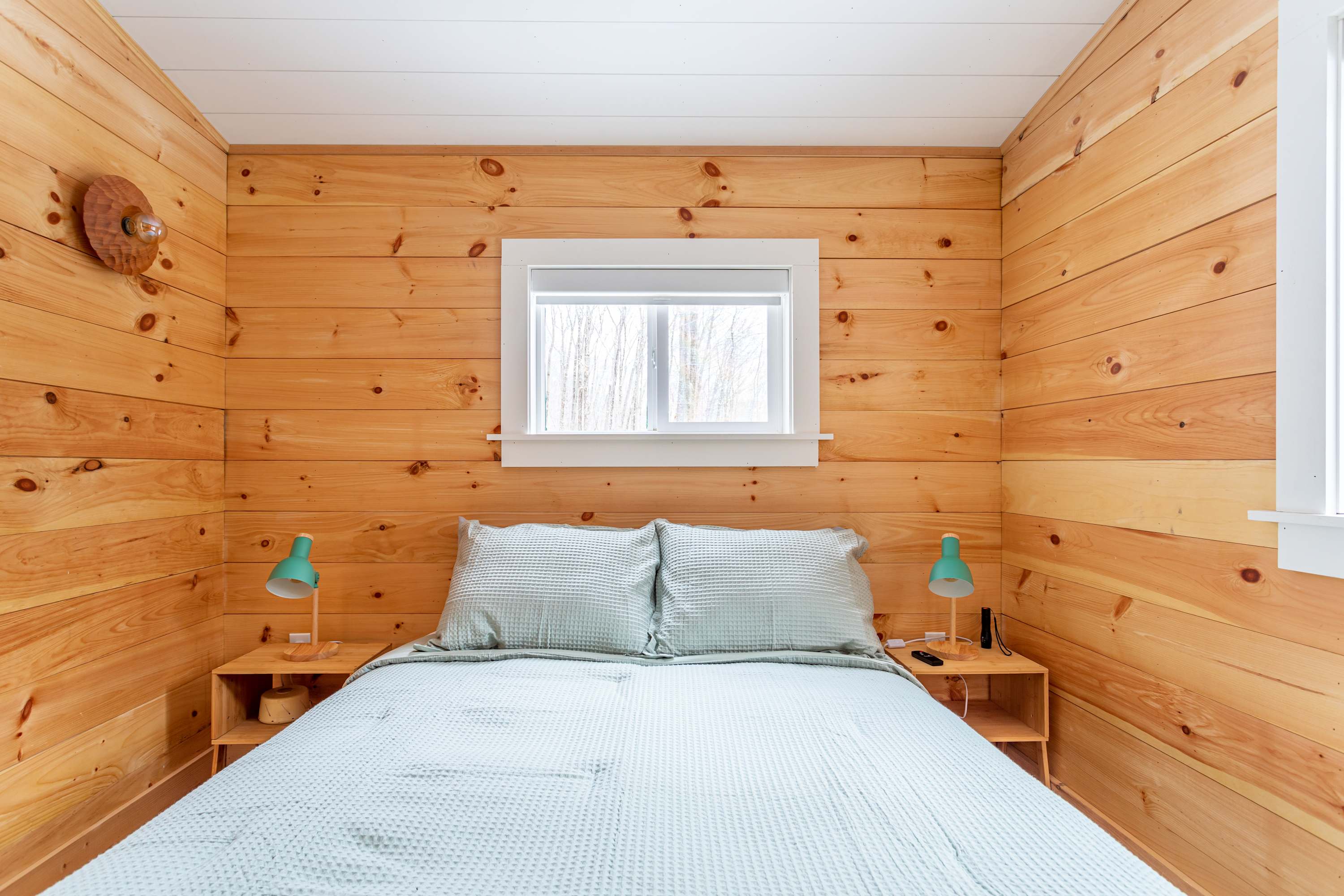 A cozy bedroom with light wood paneling on the walls and a white sloped ceiling, featuring a queen-sized bed with a light green waffle-knit comforter. Two small wooden nightstands with teal lamps flank the bed.