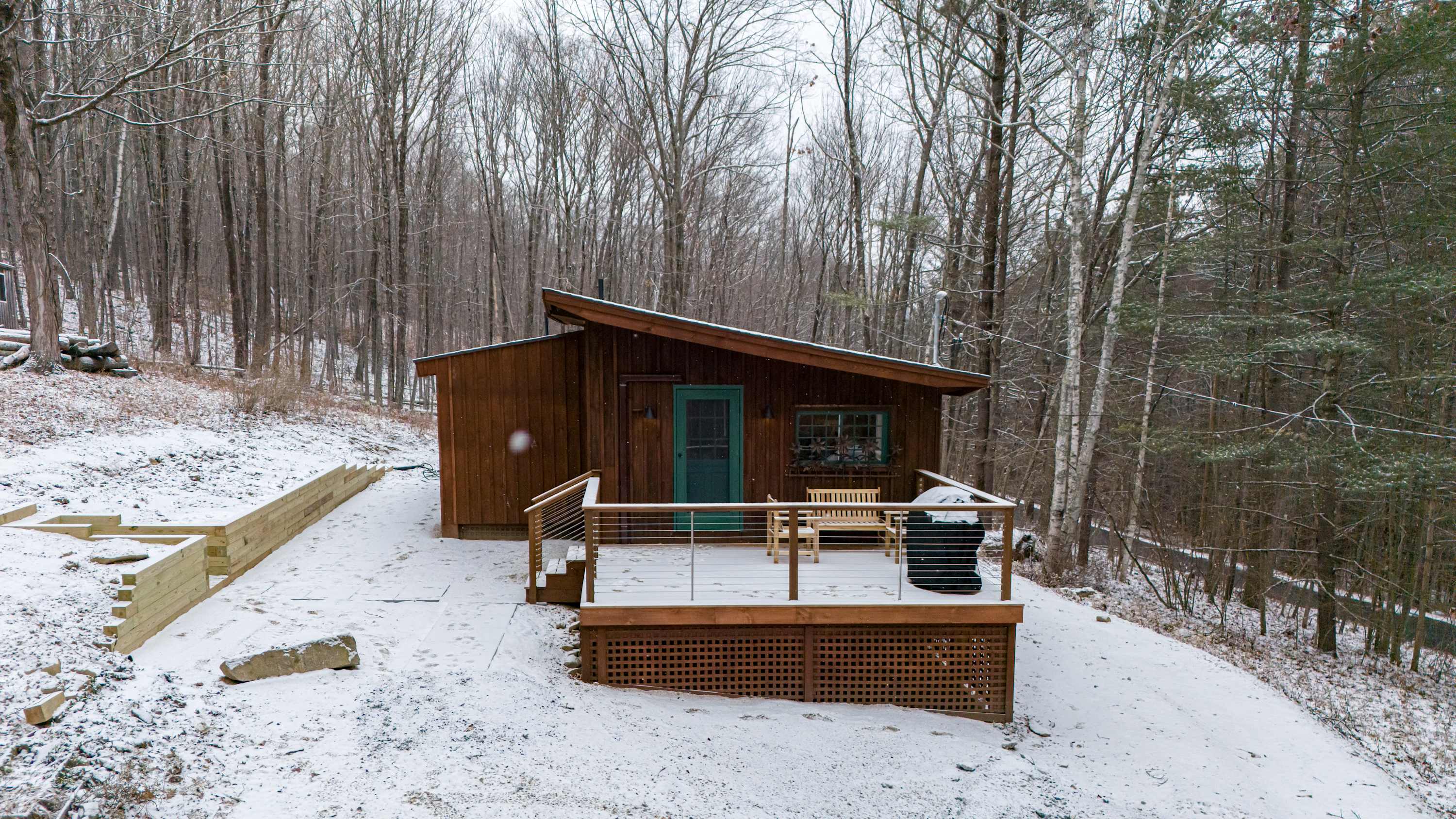 Rustic brown wooden cabin with a sloped roof and a front deck, surrounded by a dense, snow-covered forest of bare trees, showcasing its secluded setting.