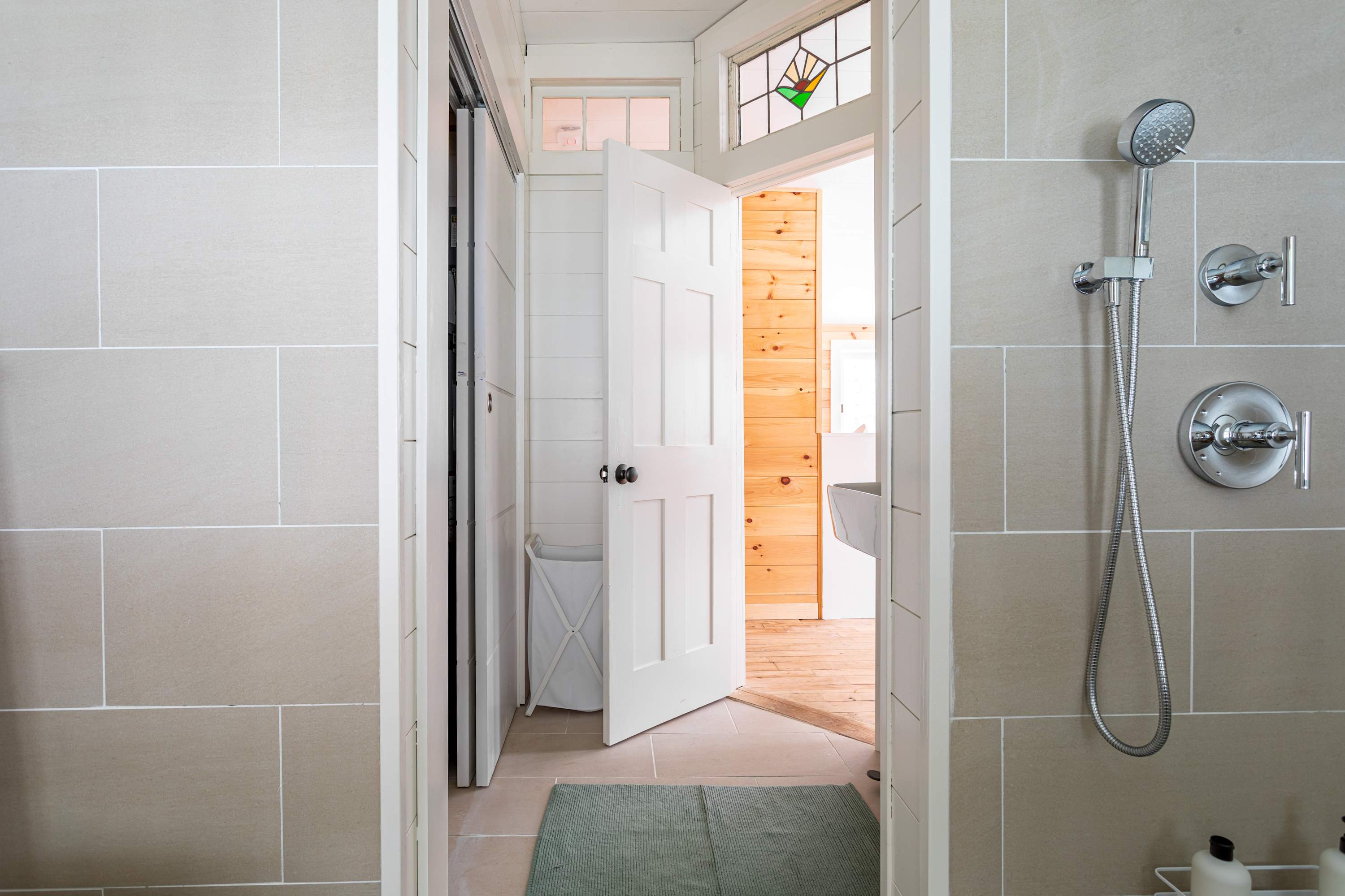 A modern shower area with light beige tiled walls, featuring a chrome shower head and controls, with a glimpse into the rest of the bathroom and a white door leading to another room.