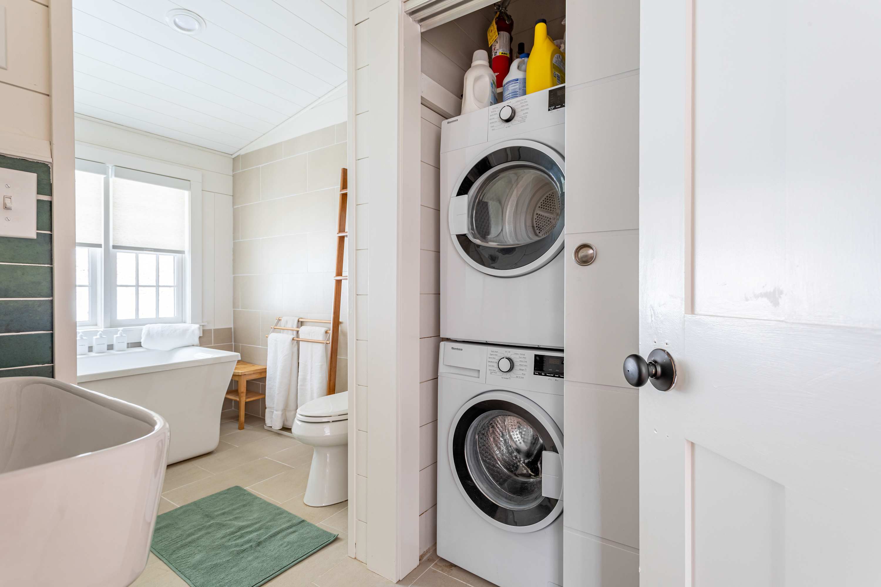 A compact laundry closet featuring a stacked white washer and dryer unit, with cleaning supplies stored on a shelf above. The closet is located within a bathroom, visible through the open door.