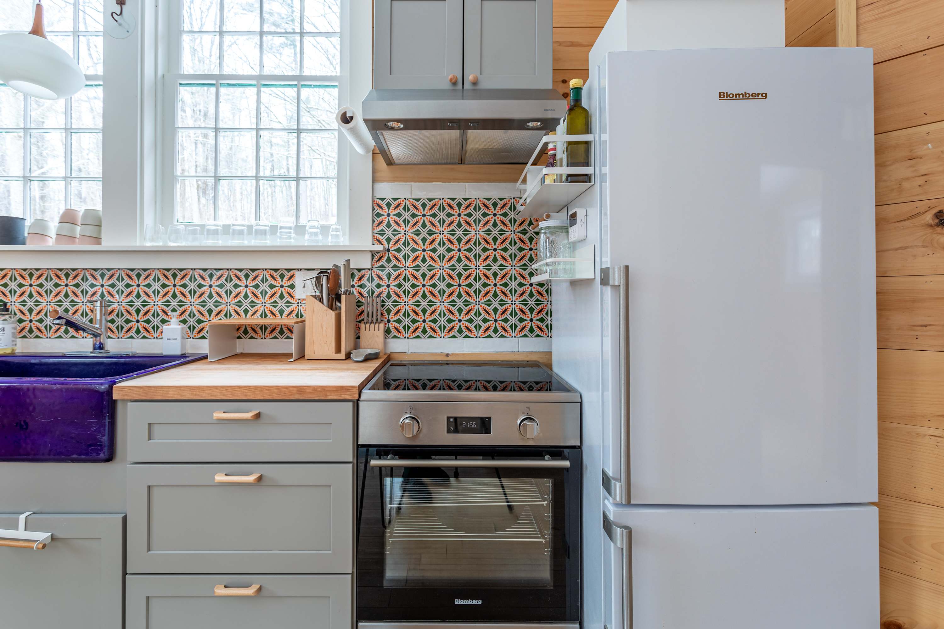 The kitchen features light wood paneling, a white Blomberg refrigerator, and a stainless steel oven with an electric cooktop. A colorful tiled backsplash adds a vibrant touch above the wood countertop.
