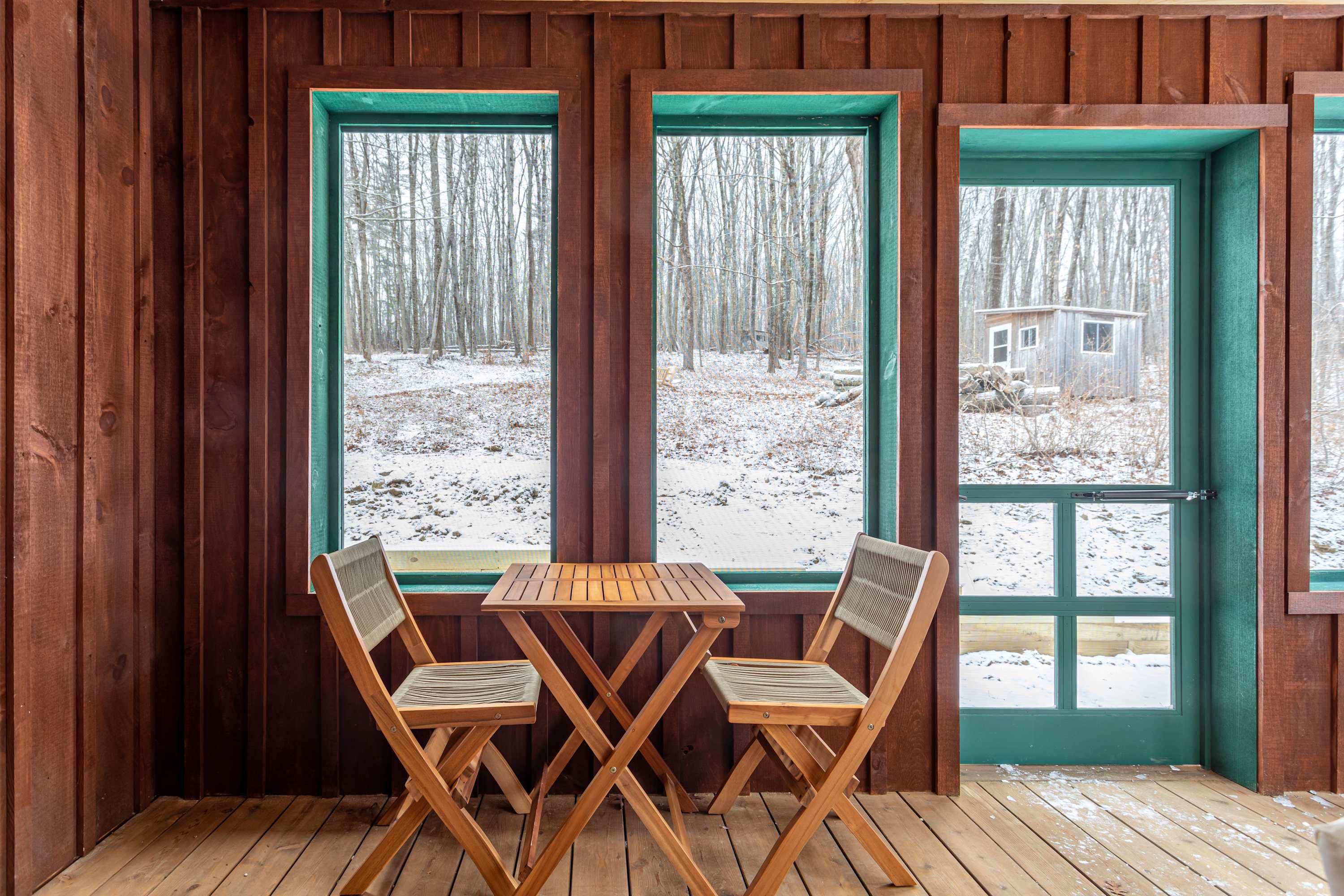 Cozy enclosed porch with dark wood paneling and teal trim around windows and a door, featuring a small wooden table and two chairs, overlooking a snow-covered forest with bare trees.