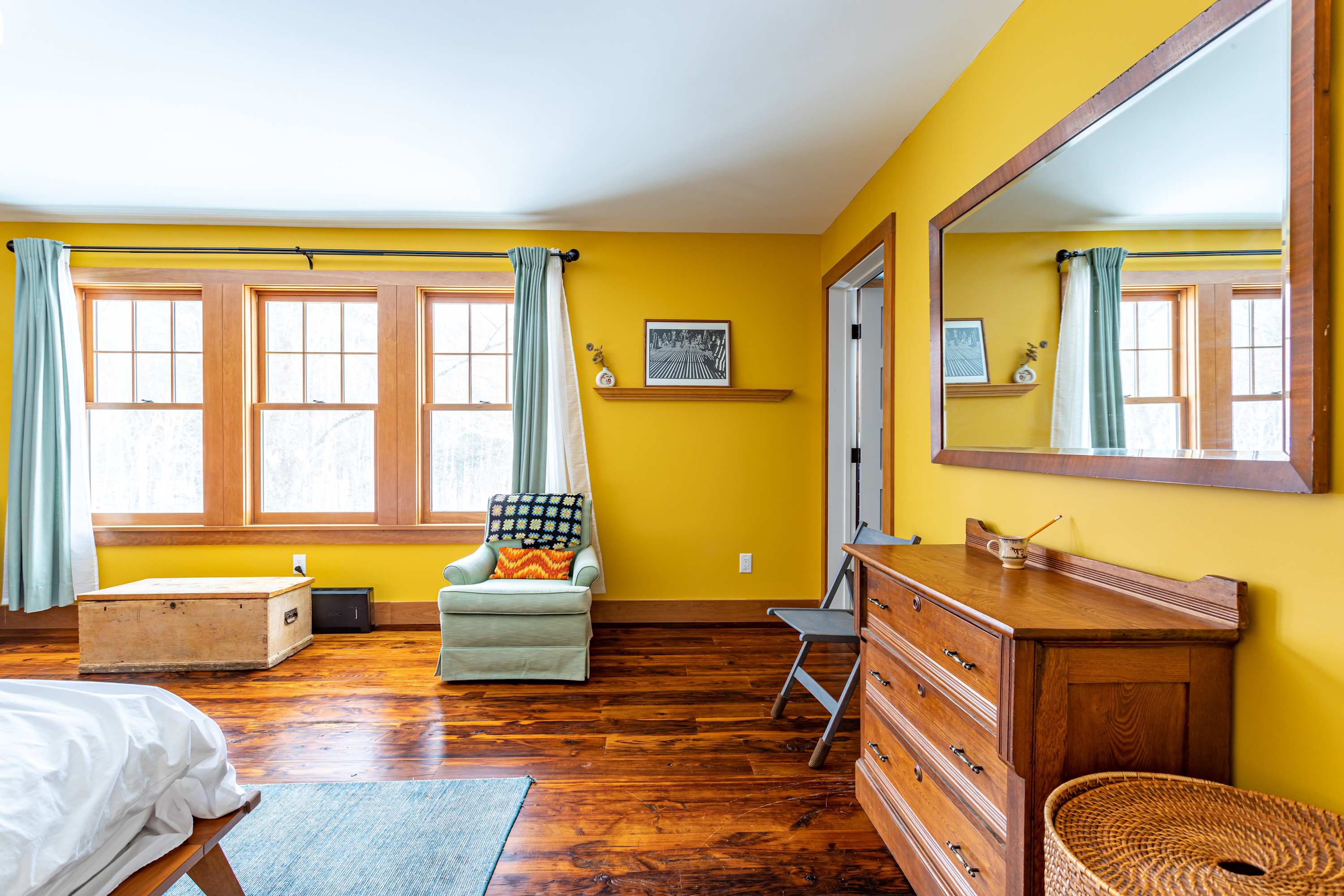 Bright bedroom with yellow walls, dark hardwood floors, and a large window with light blue curtains. A comfortable armchair and a wooden dresser with a large mirror are visible.