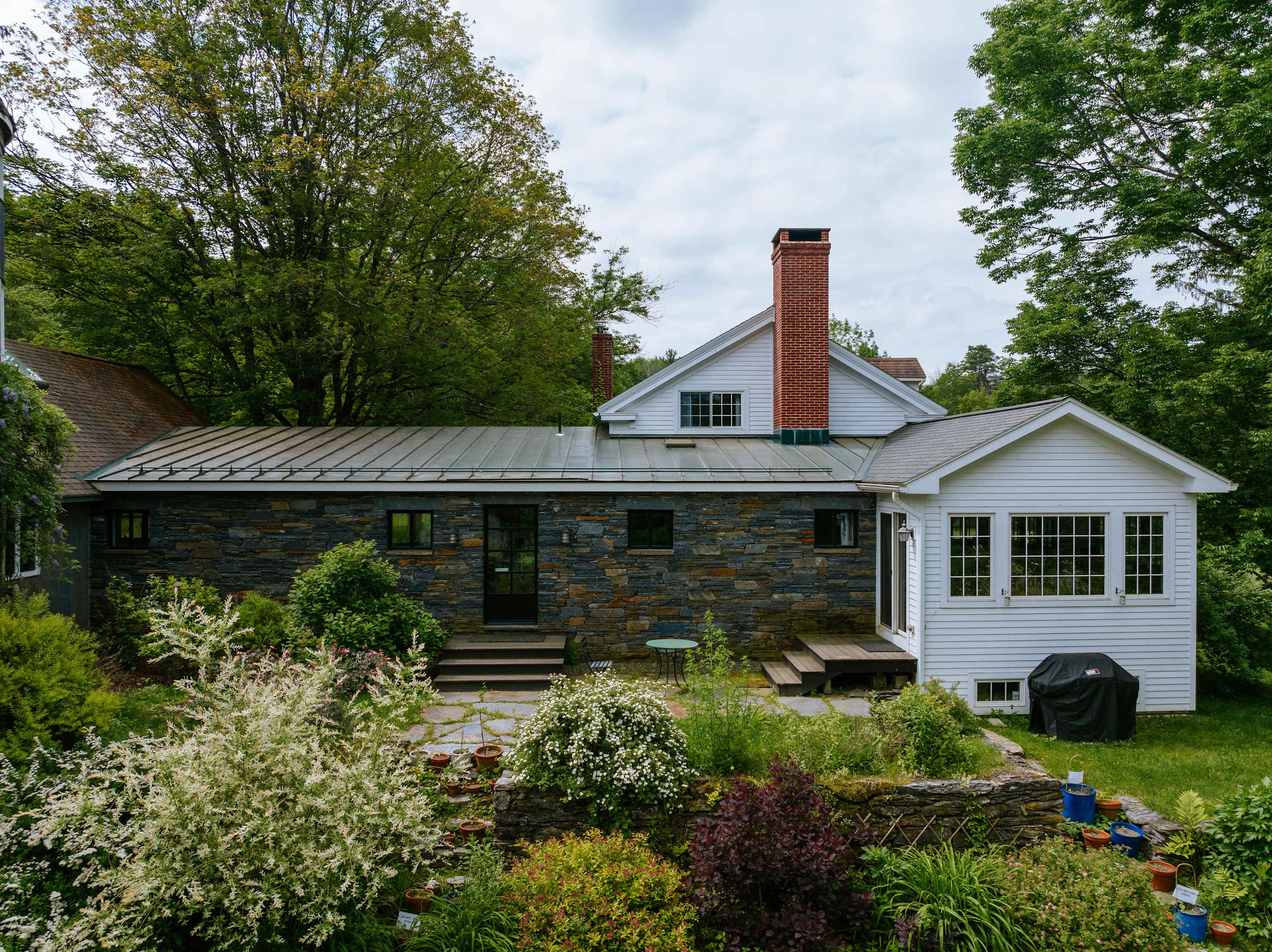 Rear view of the house featuring a stone-faced section with a metal roof, a white sunroom addition with multiple windows, and a terraced garden area with various plants and stone walls.