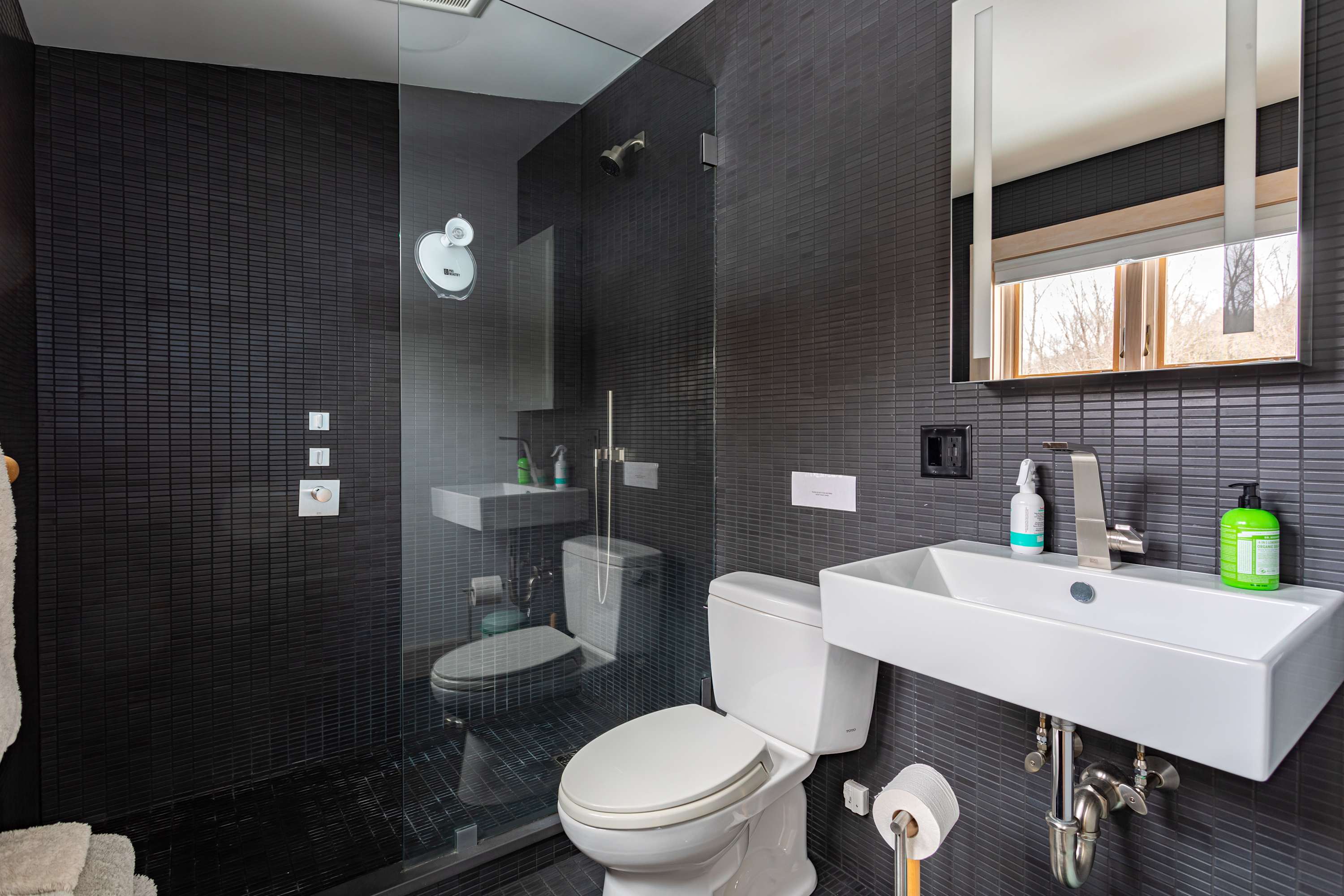 A modern bathroom features dark gray tiled walls and floor, a glass-enclosed walk-in shower, a white toilet, and a sleek wall-mounted sink with a mirror above.