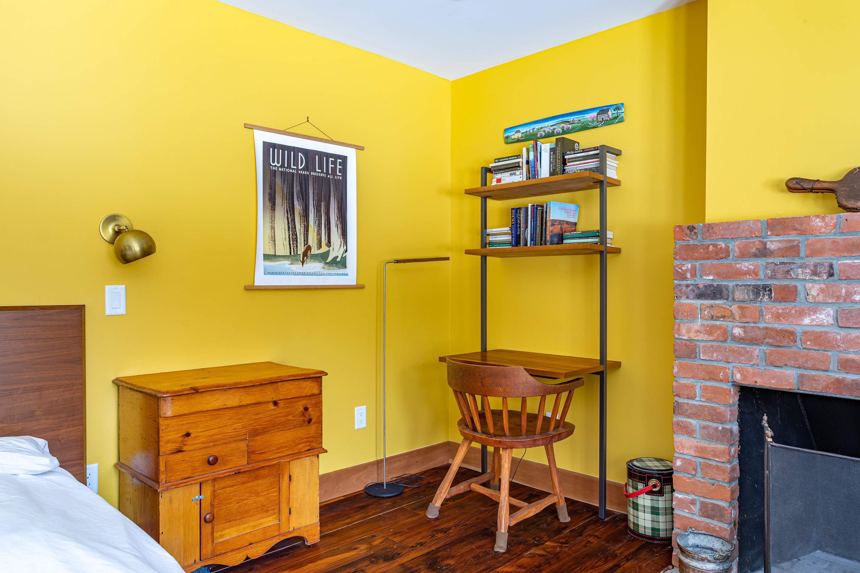 Corner of a bright yellow bedroom featuring a wooden desk with a chair, a floor lamp, and a poster on the wall. A brick fireplace is partially visible.