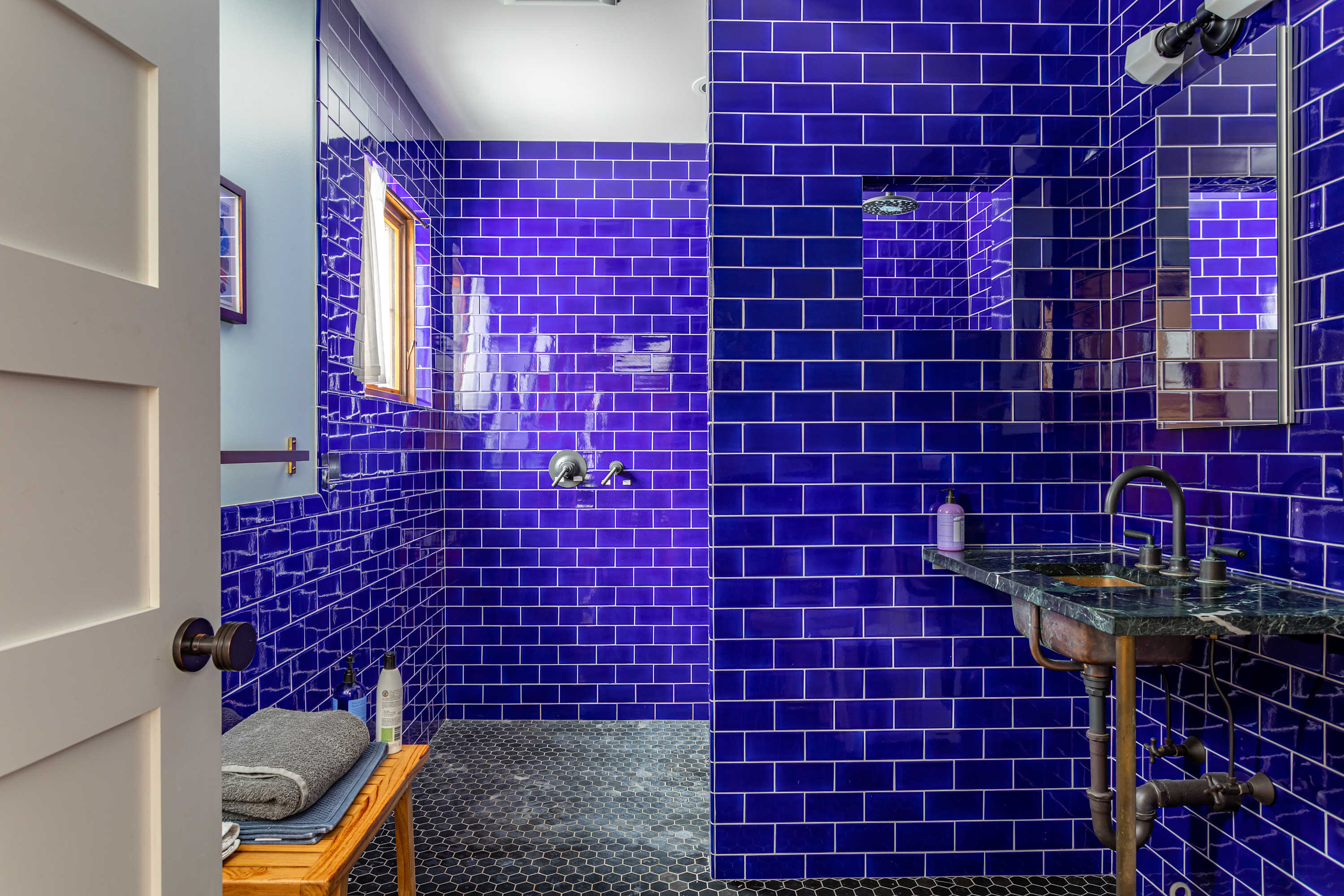 Striking bathroom featuring bright blue subway tiles covering the walls and shower area, with black hexagonal floor tiles. A unique sink with a dark marble top and exposed pipes is visible.