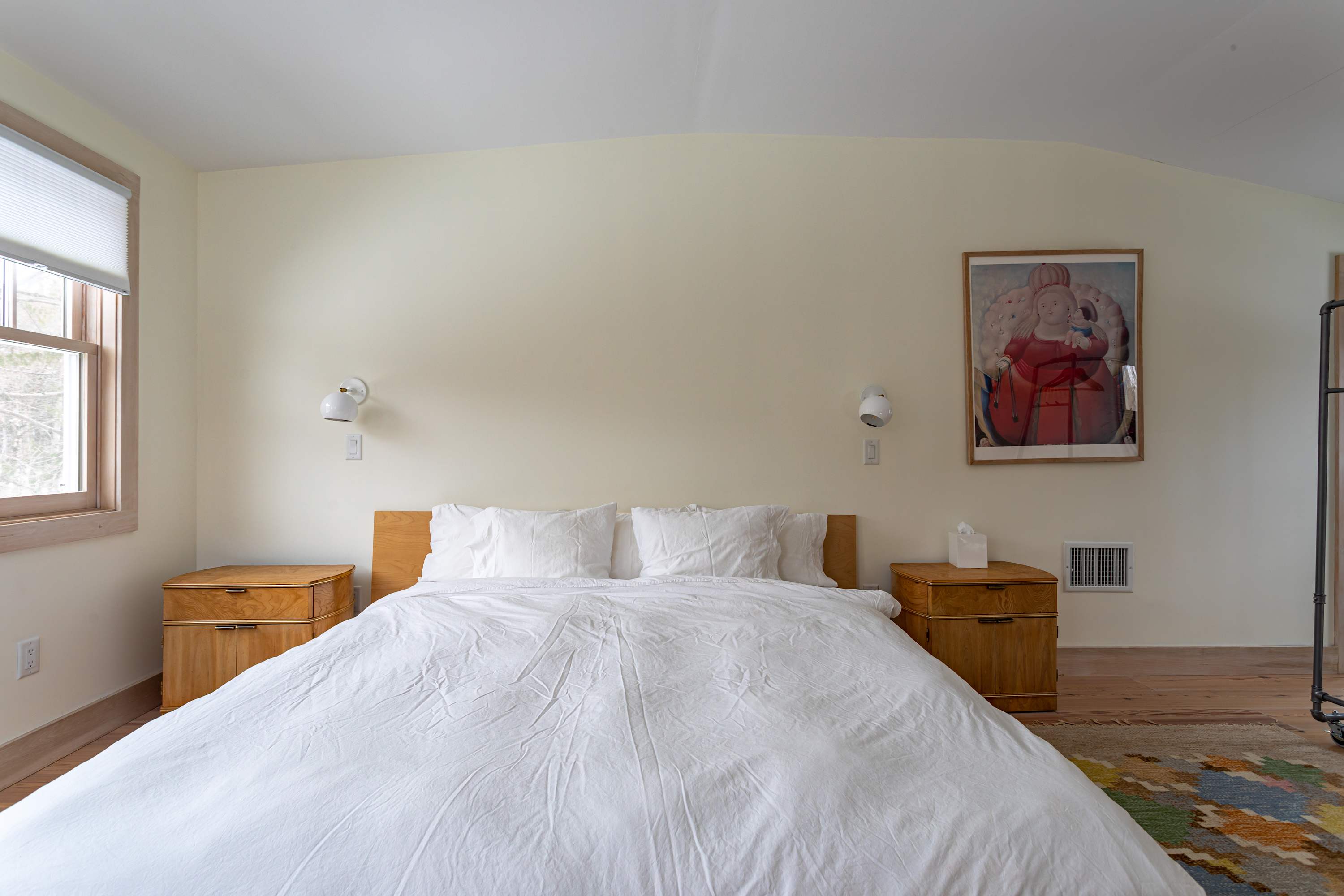 A spacious bedroom with light cream walls and hardwood floors features a modern wooden bed frame with white bedding, flanked by wooden nightstands with integrated lighting. A colorful abstract rug adds warmth.