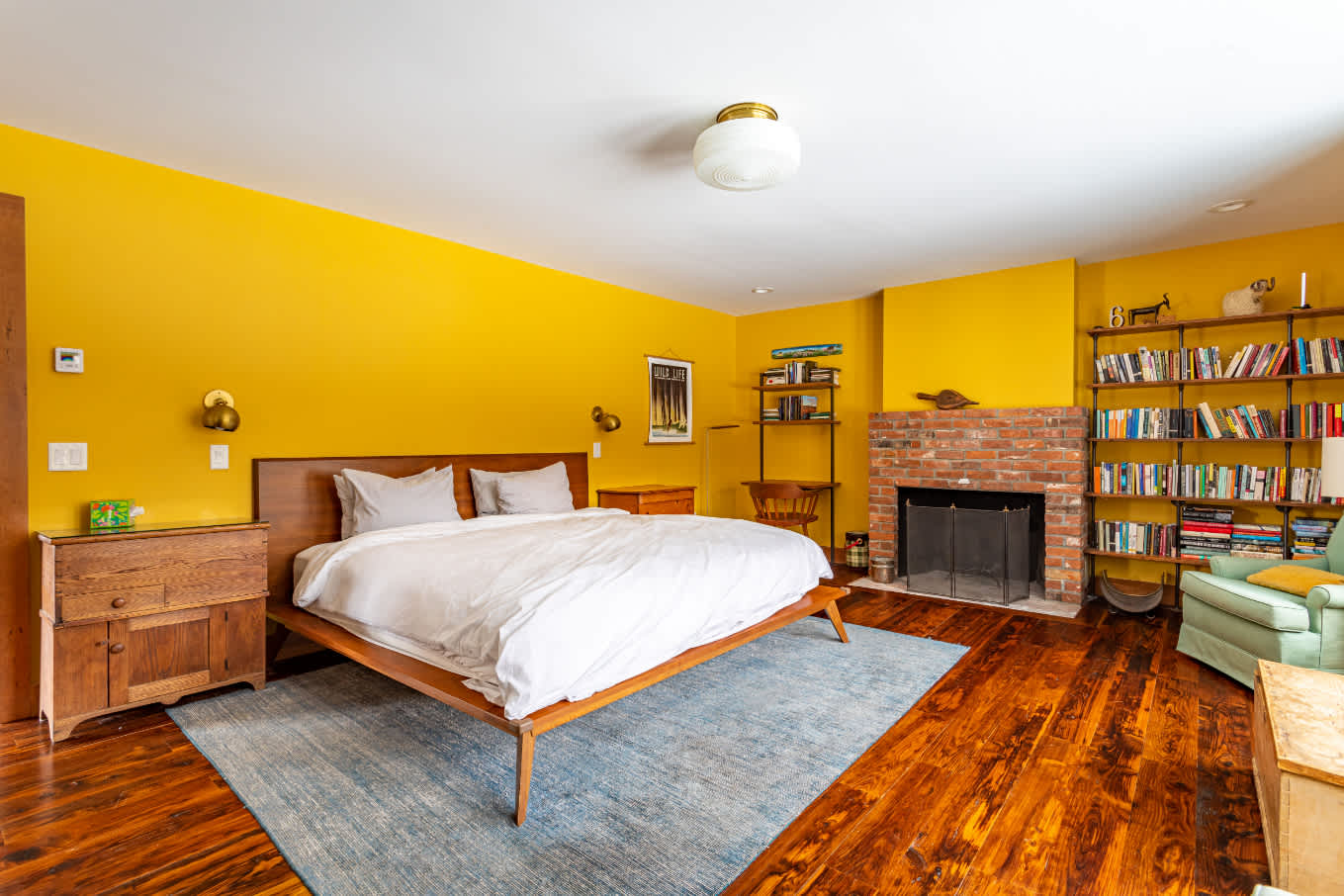 Spacious bedroom with vibrant yellow walls, dark hardwood floors, and a modern wooden bed frame with white bedding. A brick fireplace and built-in bookshelves line one wall.