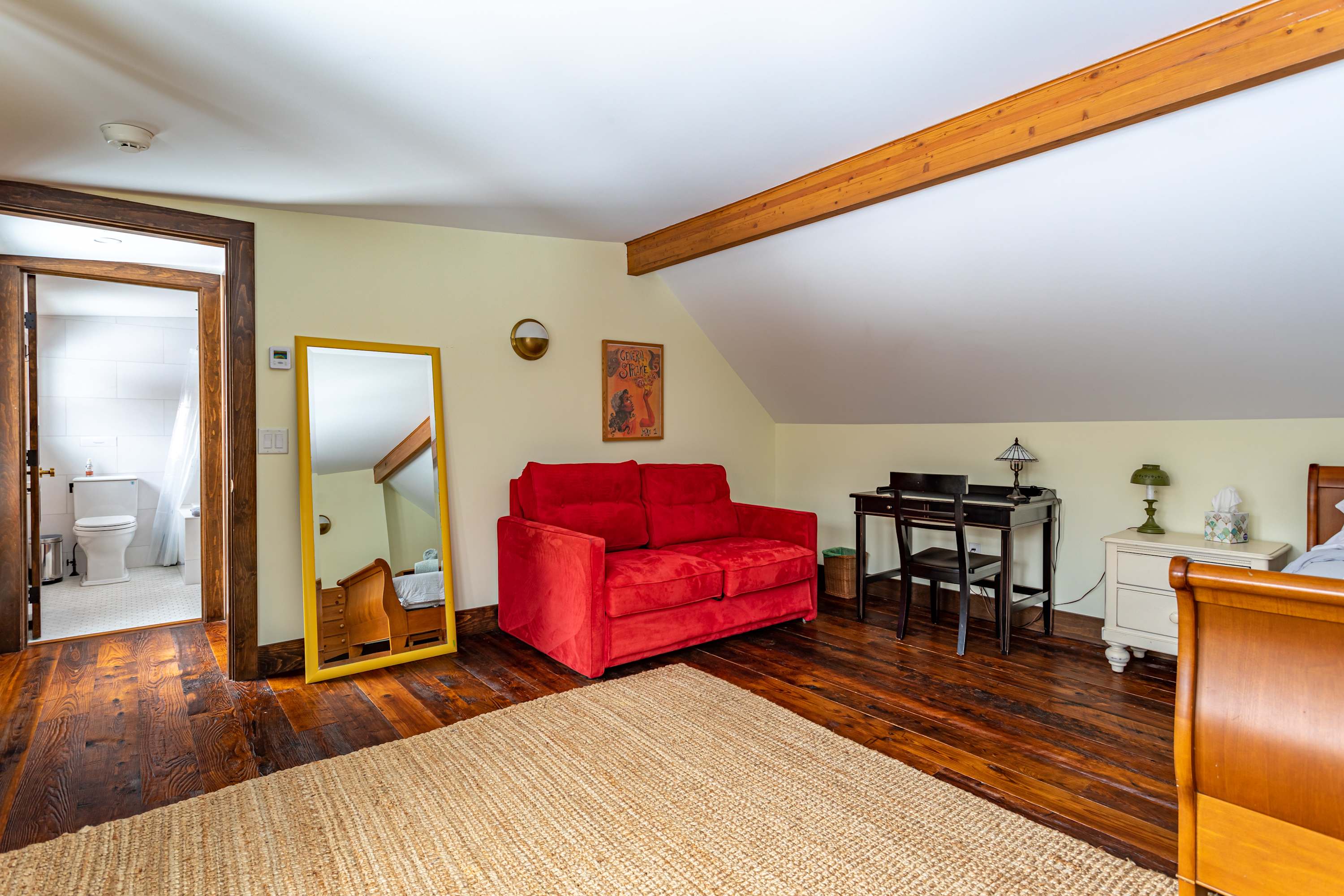 Attic bedroom with dark hardwood floors and light yellow walls, featuring a red two-seater sofa, a black desk, and a full-length mirror. A doorway leads to a white-tiled bathroom.