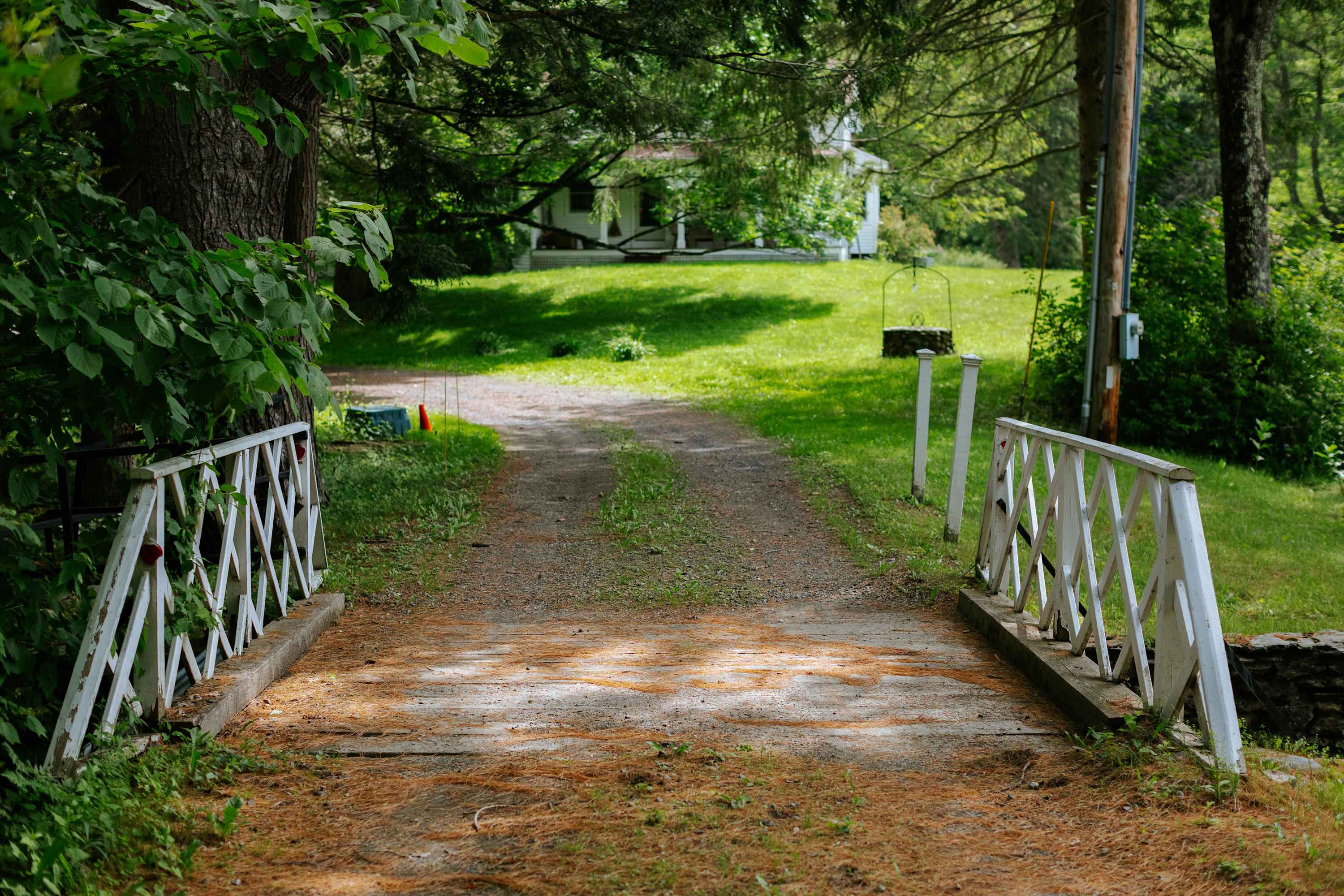 A gravel driveway leads over a small white wooden bridge with lattice railings, towards a white house partially visible through dense green trees in the background. The path is surrounded by green grass and pine needles.
