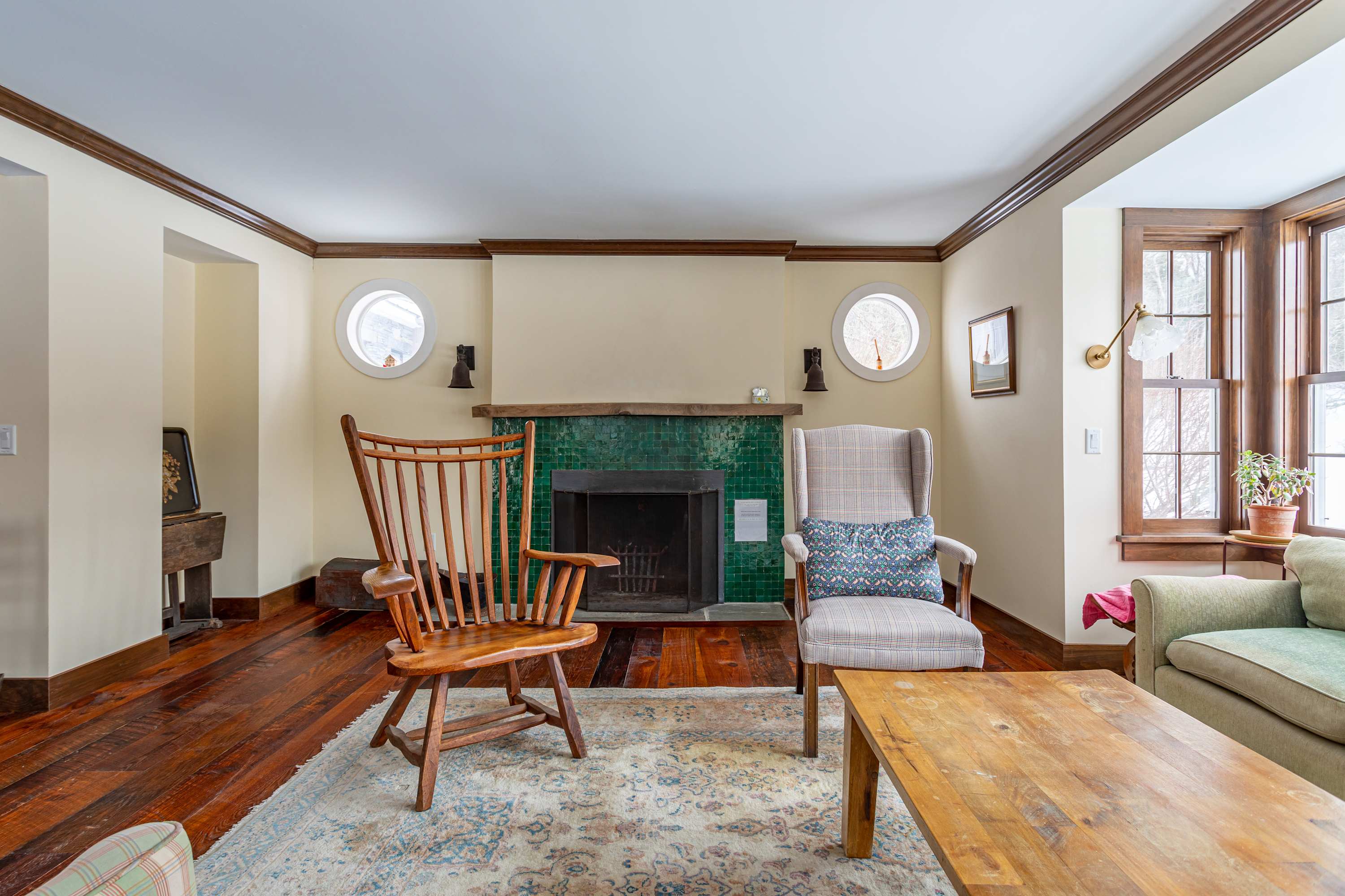 Cozy living room with dark hardwood floors, a patterned area rug, and a fireplace with green tiled surround, flanked by two round windows and two armchairs.