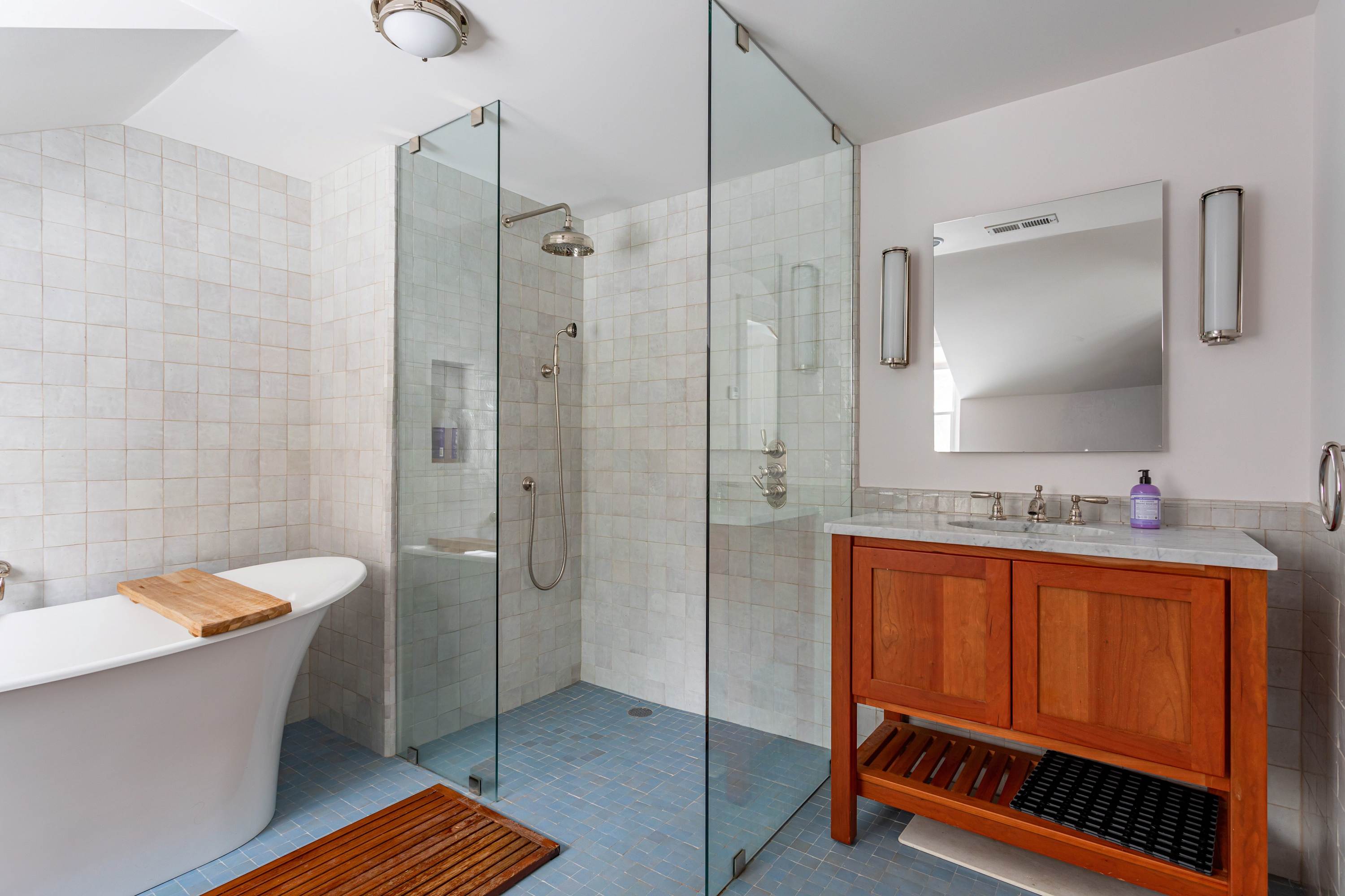 Modern bathroom with light blue floor tiles and white square wall tiles, featuring a glass-enclosed shower, a freestanding tub, and a wooden vanity with a marble top and mirror.