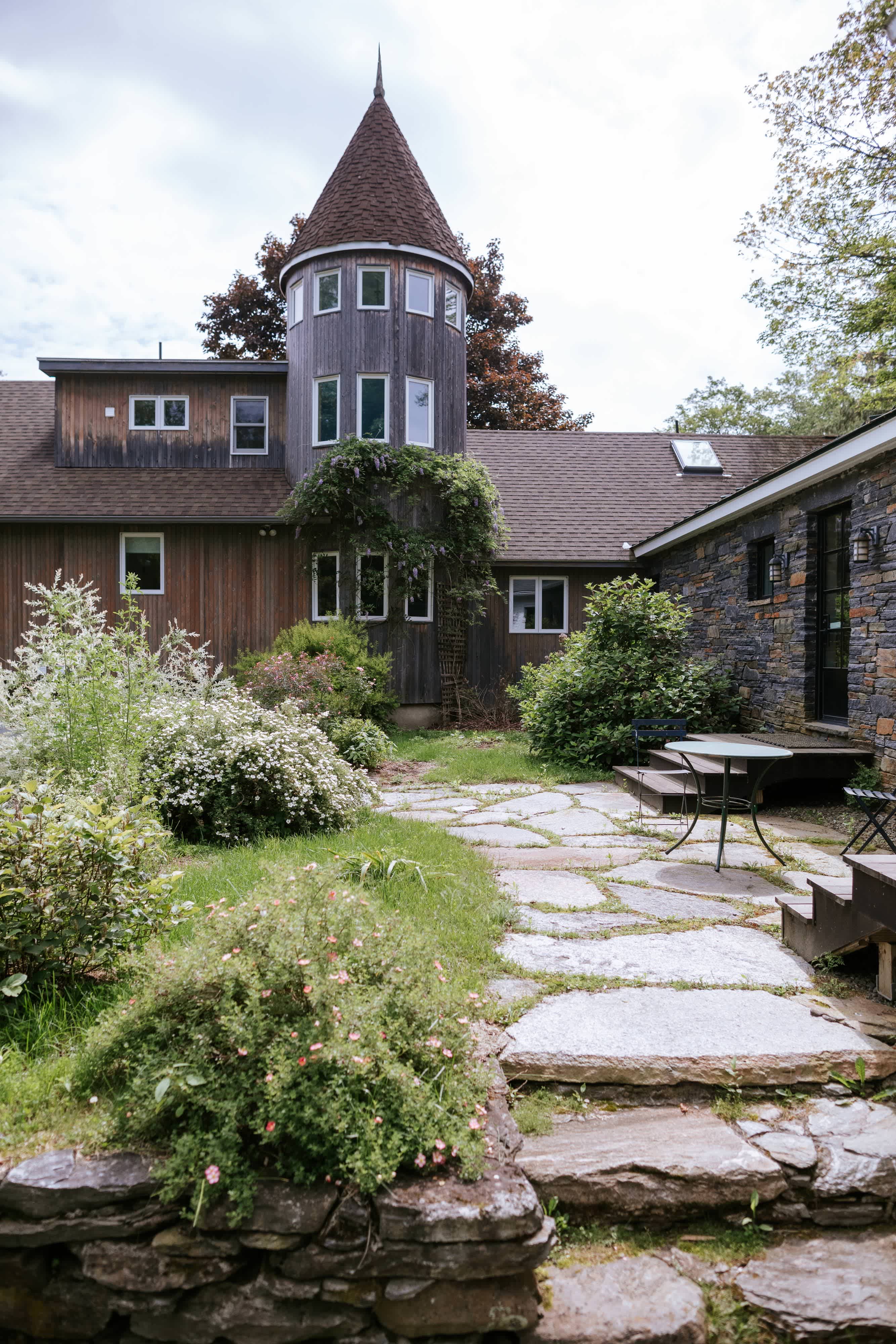 The back of the house features a unique round tower with multiple windows and a conical roof, connected to a stone-sided section with large windows and a wooden-sided section. A stone patio with outdoor furniture is surrounded by lush landscaping.