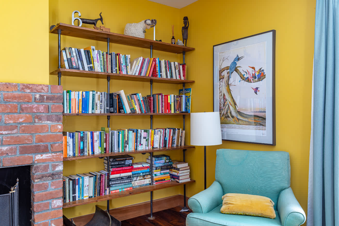 Cozy corner of a yellow bedroom with a brick fireplace, built-in wooden bookshelves filled with books, and a light blue armchair with a floor lamp.