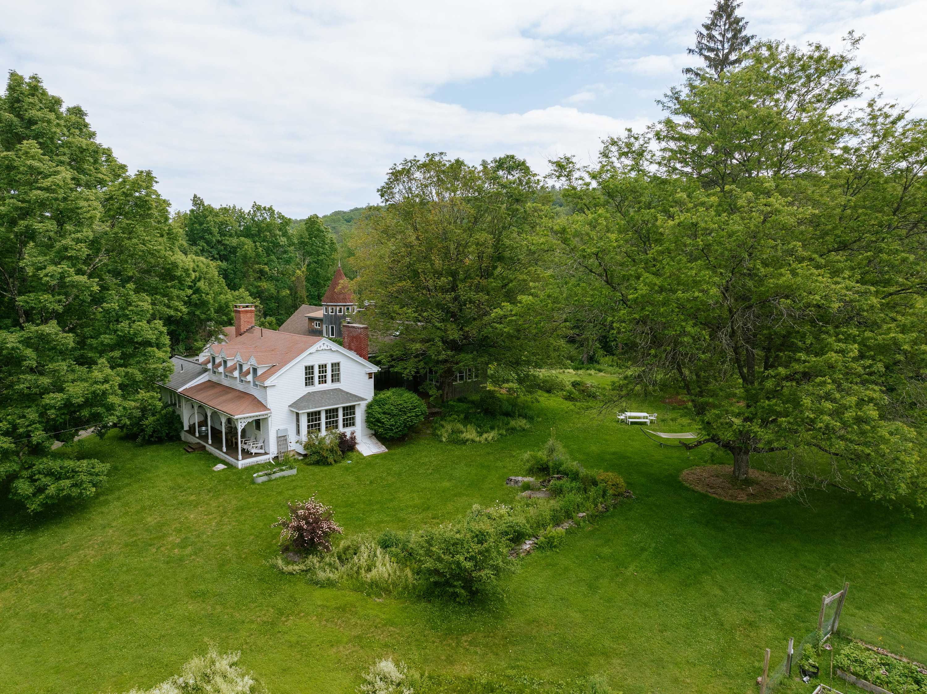 Expansive aerial view of the white, traditional-style house with a red roof, surrounded by a vast, manicured green lawn and dense mature trees, under a partly cloudy sky.