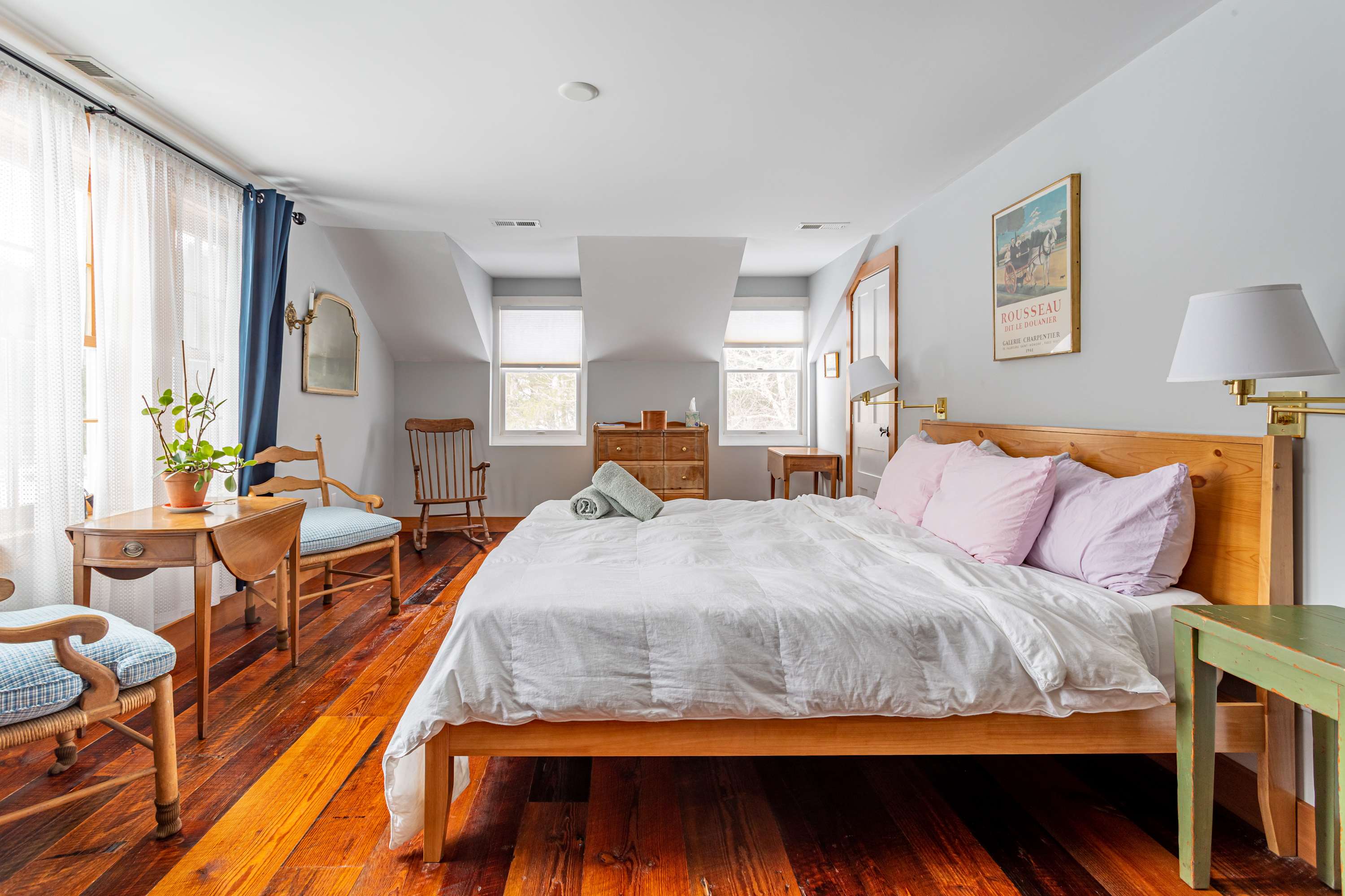 Spacious bedroom with warm-toned hardwood floors, a light wood bed frame with white and pink bedding, a wooden dresser, and a small desk with a chair, under sloped ceilings.