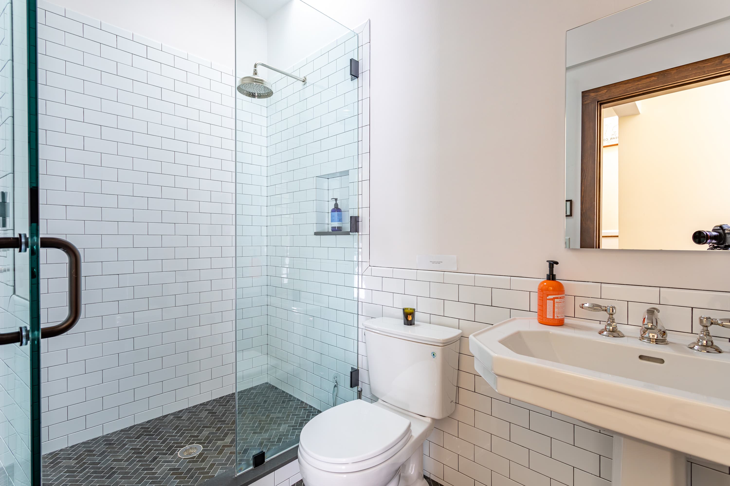 Modern bathroom with white subway tile walls, a glass-enclosed shower with dark gray herringbone floor tiles, a white toilet, and a pedestal sink with a mirror above.