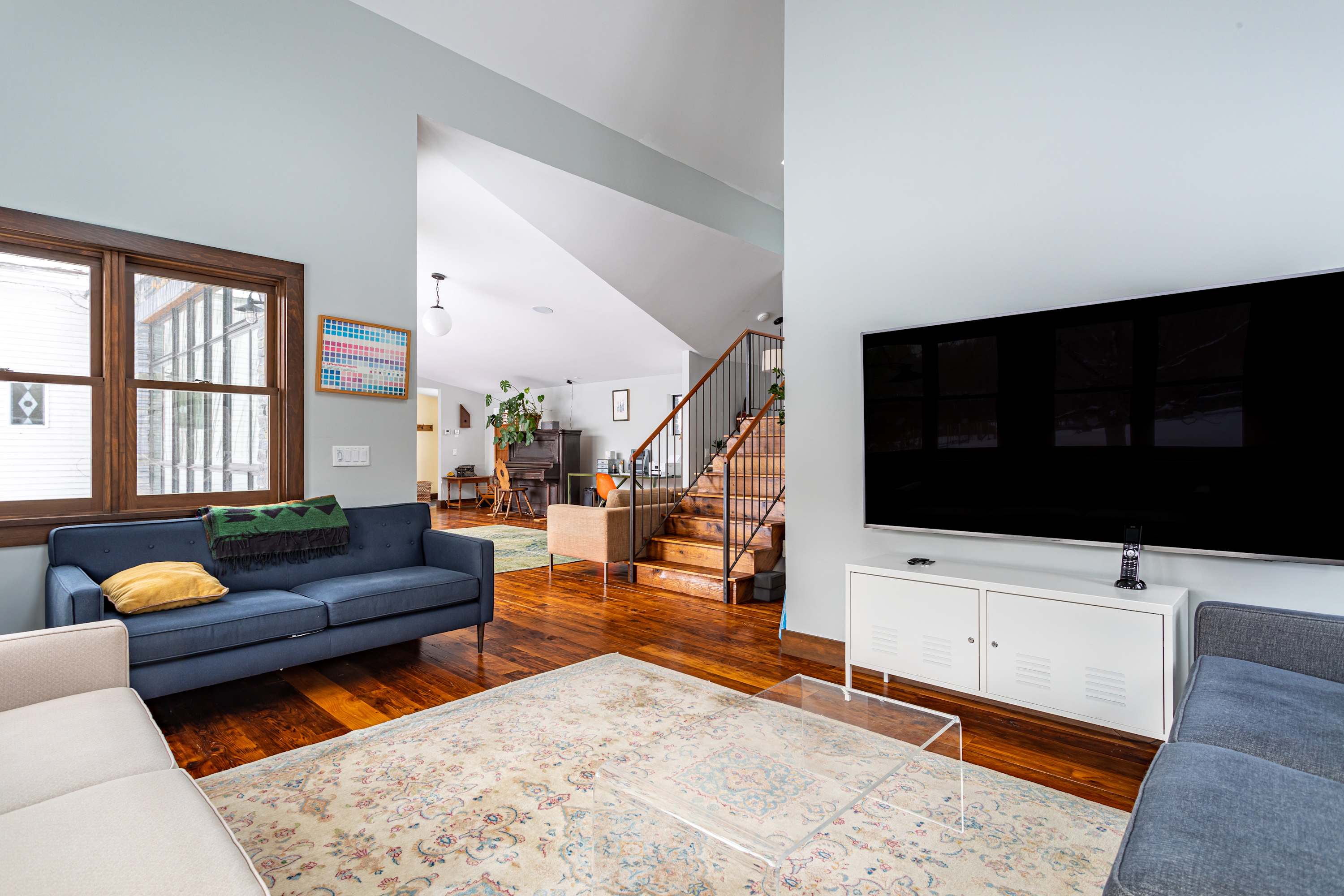 Modern living room with light blue walls, a blue sofa, a large wall-mounted TV above a white console, and a staircase leading upstairs in the background, all on polished hardwood floors.