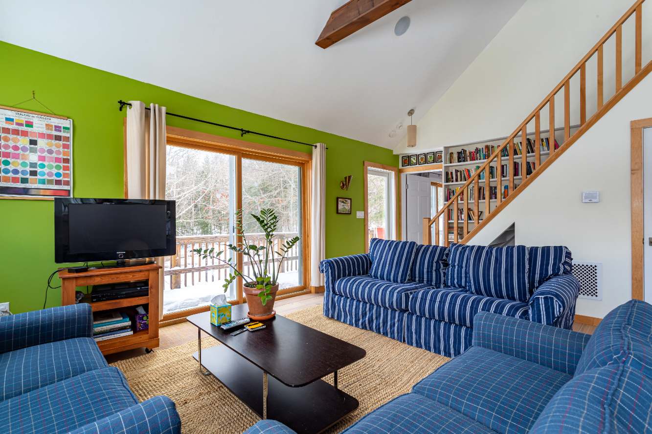 A vibrant living room features lime green walls, a blue striped sofa, and a blue plaid armchair on a jute rug, with large glass doors opening to a snowy deck. A wooden staircase with built-in bookshelves leads upstairs.