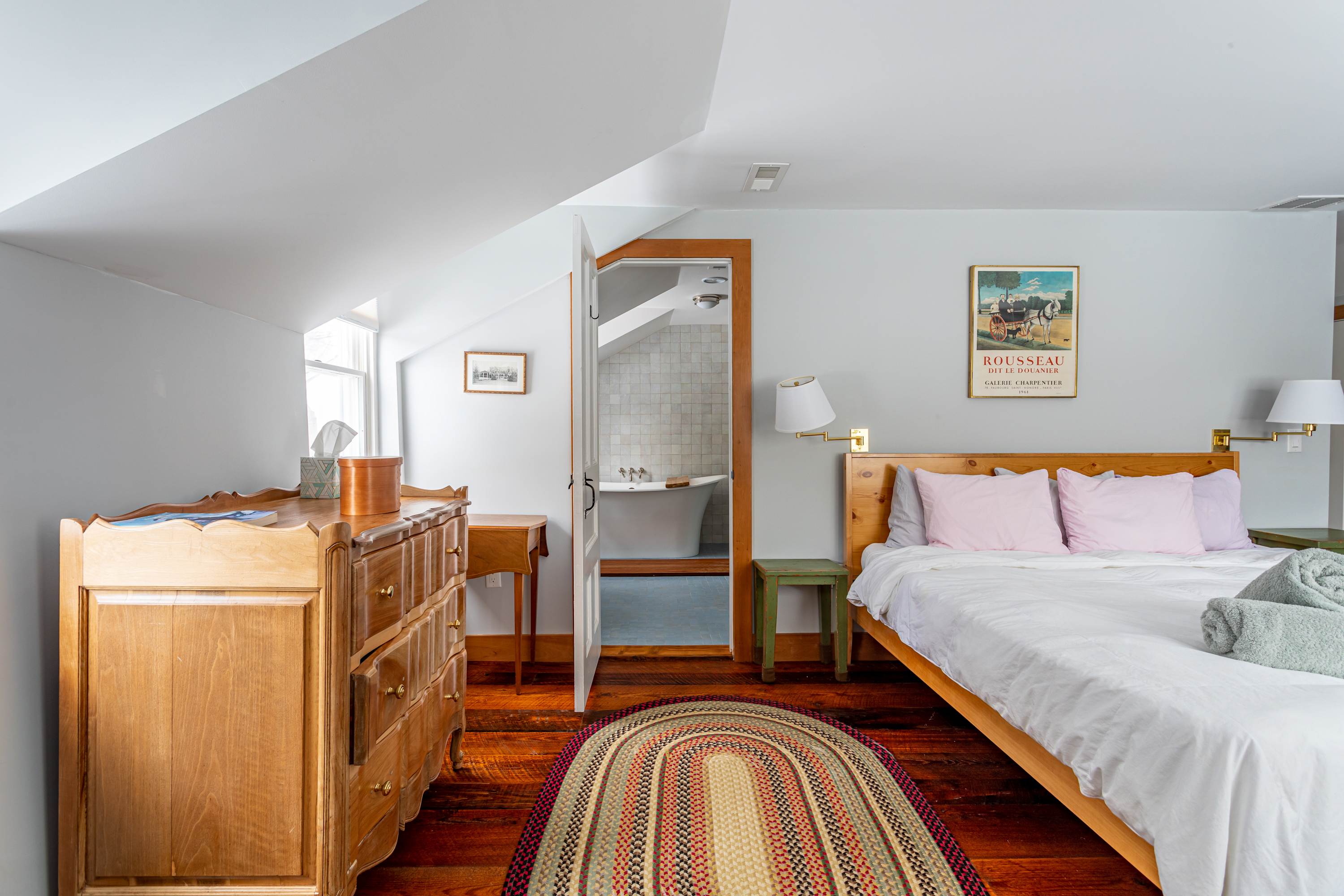 View of the bedroom with warm hardwood floors and a patterned rug, showing a light wood bed, a dresser, and an open doorway leading to an en-suite bathroom with a freestanding tub.
